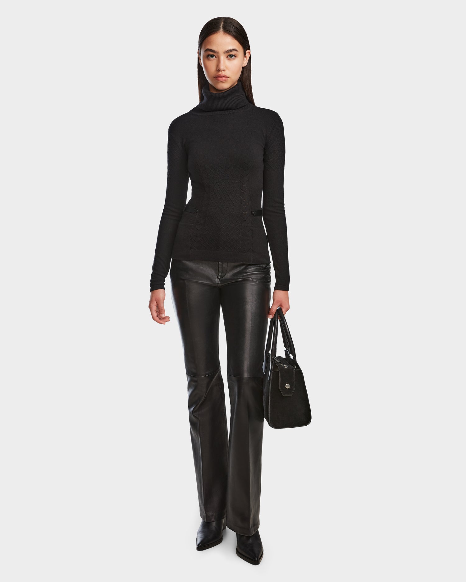 A woman faces forward against a plain white background, wearing the Rudsak LAURA - 8524596 Black womens knit turtleneck sweater, black leather pants with vegan leather trim, pointed-toe shoes, and holding a black handbag in her left hand.