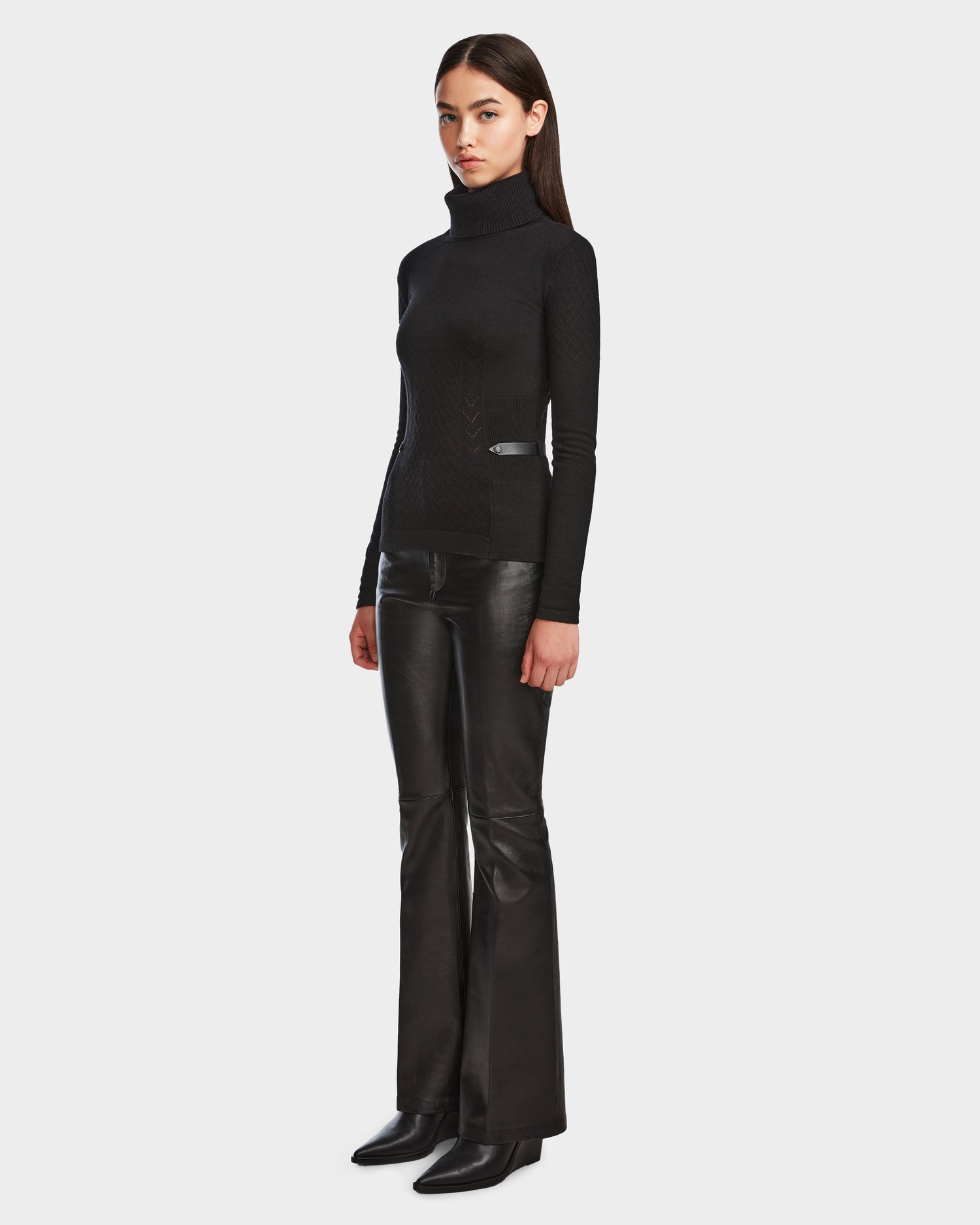 A woman with straight dark hair and a neutral expression stands against a plain background wearing the Rudsak LAURA - 8524596 Black leather flared pants with vegan leather trim, paired with a knit turtleneck sweater and black heeled boots.