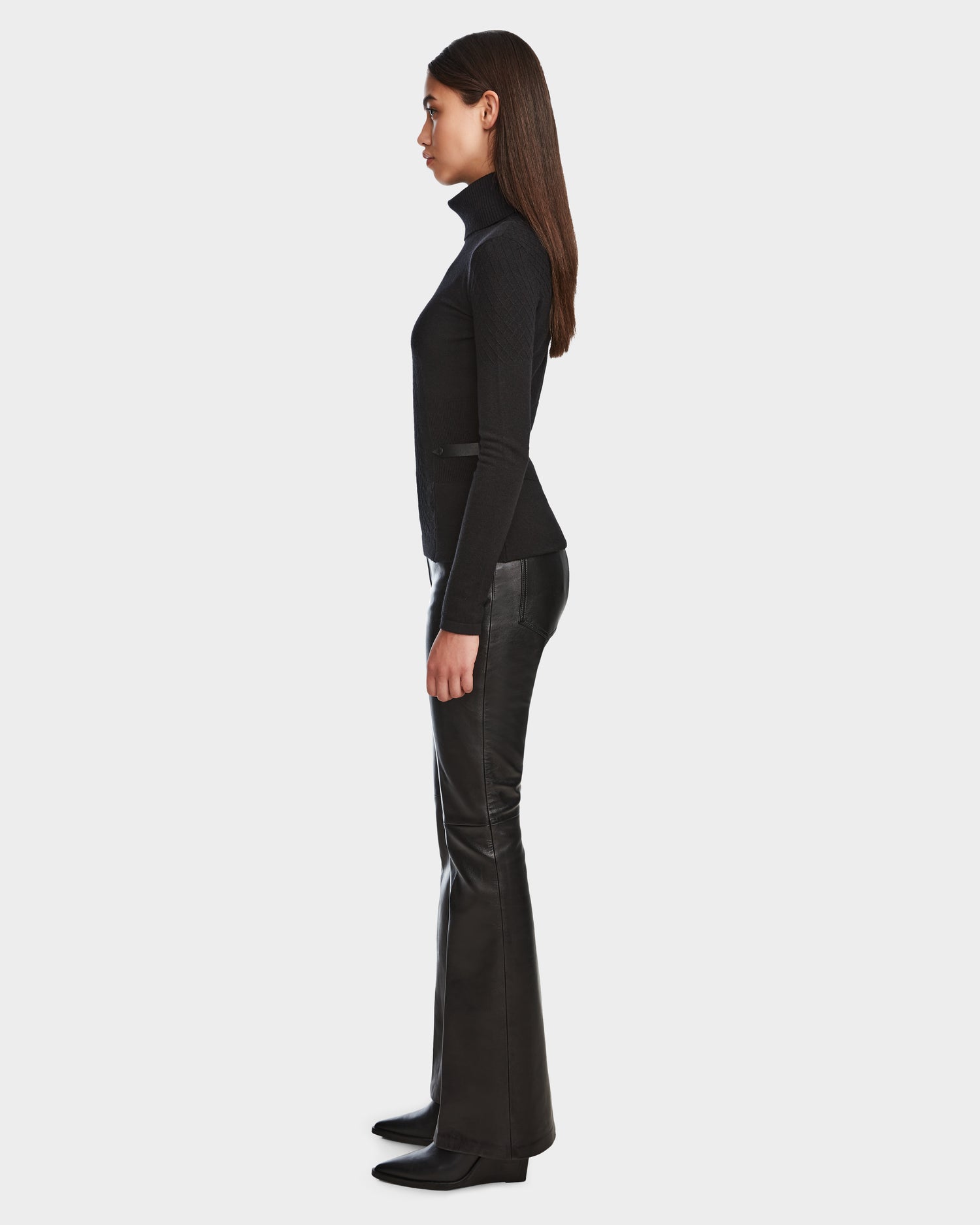 A woman in profile wears the LAURA - 8524596 Black knit turtleneck sweater by Rudsak with black flared leather pants and heeled boots, standing against a plain white background.