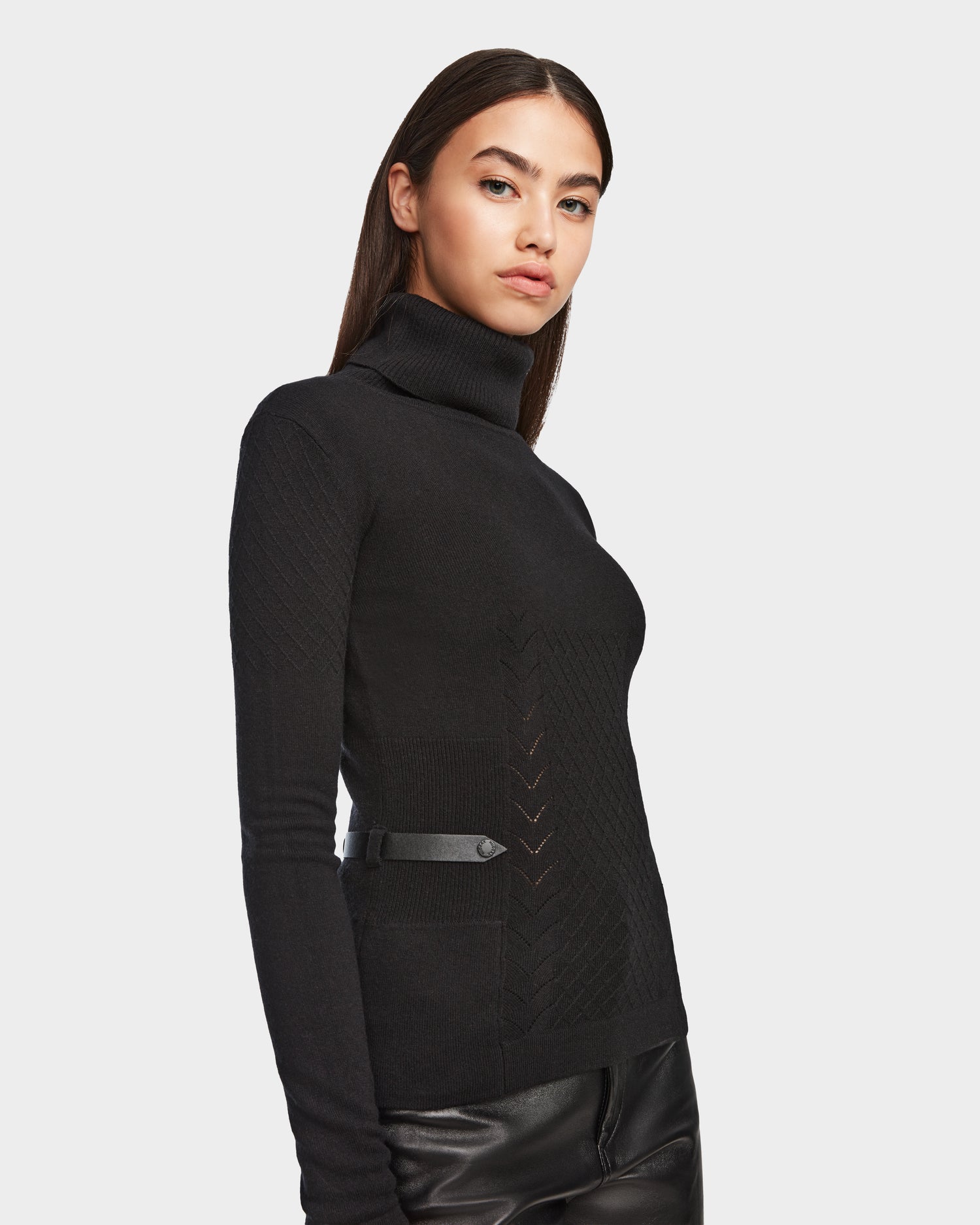 A woman with long dark hair models the Rudsak LAURA - 8524596 Black fitted women’s knit turtleneck sweater featuring subtle chevron patterns, paired with black leather pants against a plain light background.
