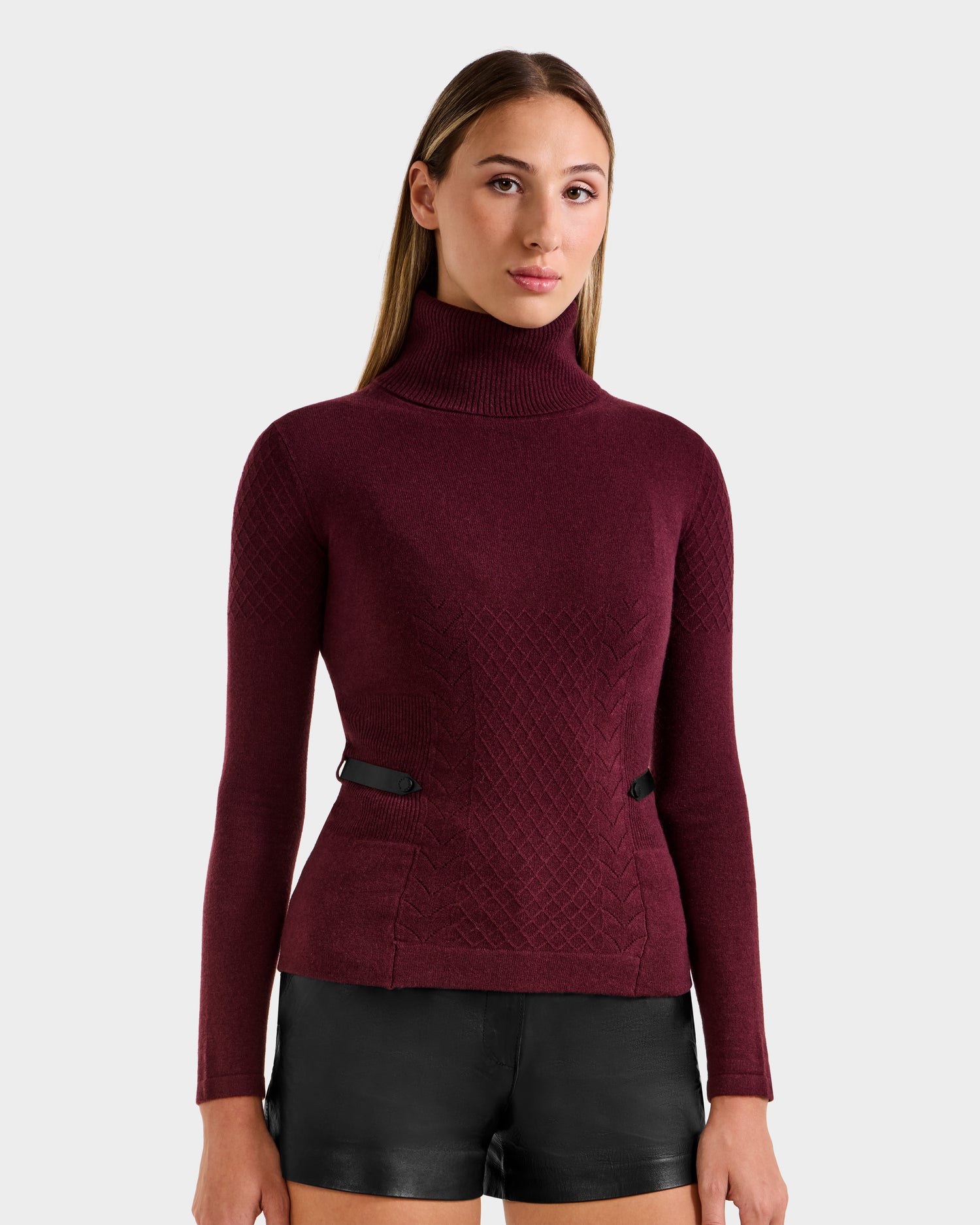 A woman with straight, light brown hair wears the Rudsak LAURA - 8524596 Bordeaux women's knit turtleneck sweater paired with black leather shorts, standing against a plain light gray background.