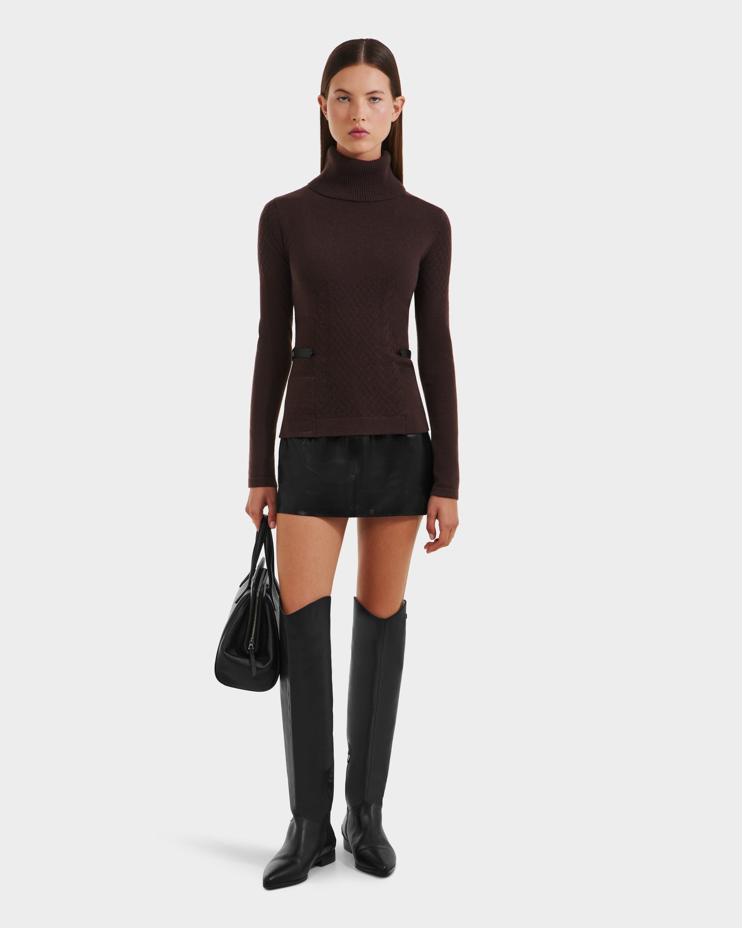 A woman poses against a plain background wearing the Rudsak LAURA - 8524596 Mocha fitted womens knit turtleneck sweater, paired with a short black skirt, black knee-high boots, and a black handbag by her side.