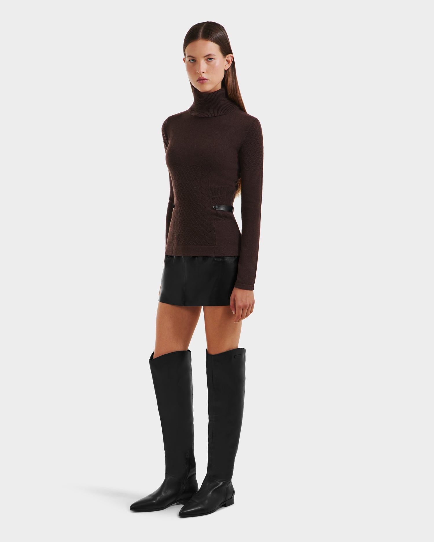 A woman with straight brown hair and a neutral expression wears the Rudsak LAURA - 8524596 Mocha women’s knit turtleneck sweater, paired with a black mini skirt and tall black boots against a plain background.