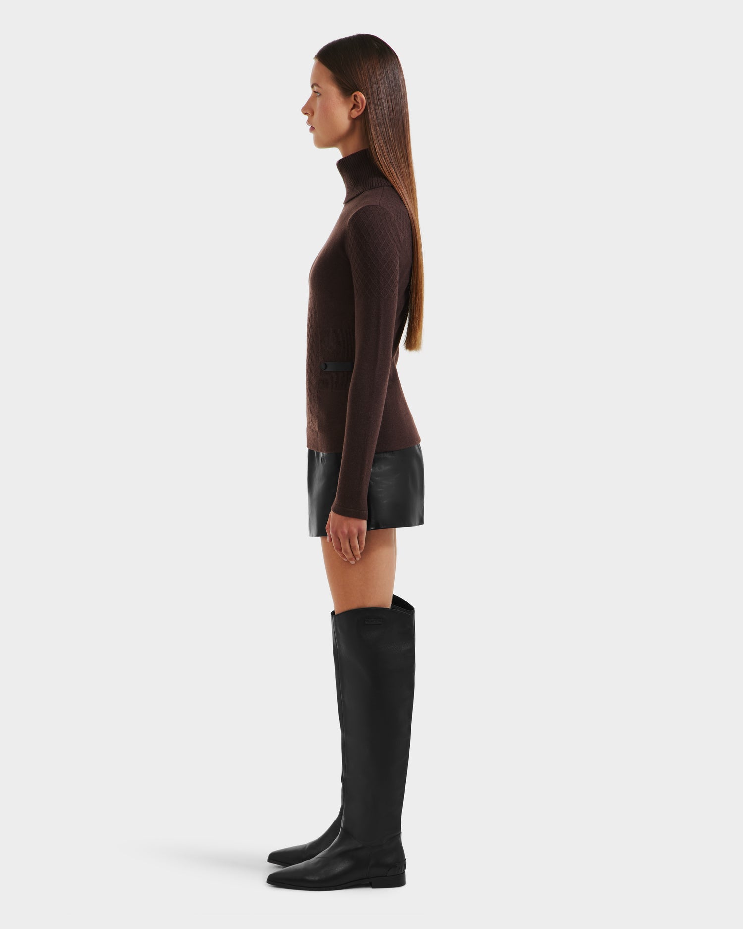 A woman in profile stands against a white background, wearing the Rudsak LAURA - 8524596 Mocha knit turtleneck sweater, paired with a black leather mini skirt and knee-high black boots. Her straight brown hair is worn down.