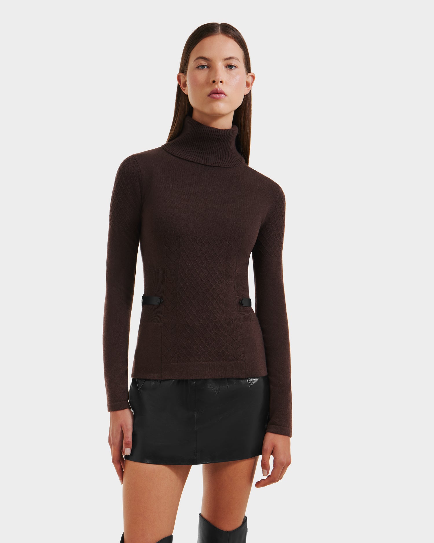 A young woman with straight brown hair wears the Rudsak LAURA - 8524596 Mocha women’s knit turtleneck sweater and a black mini skirt with vegan leather trim, standing against a plain white background.