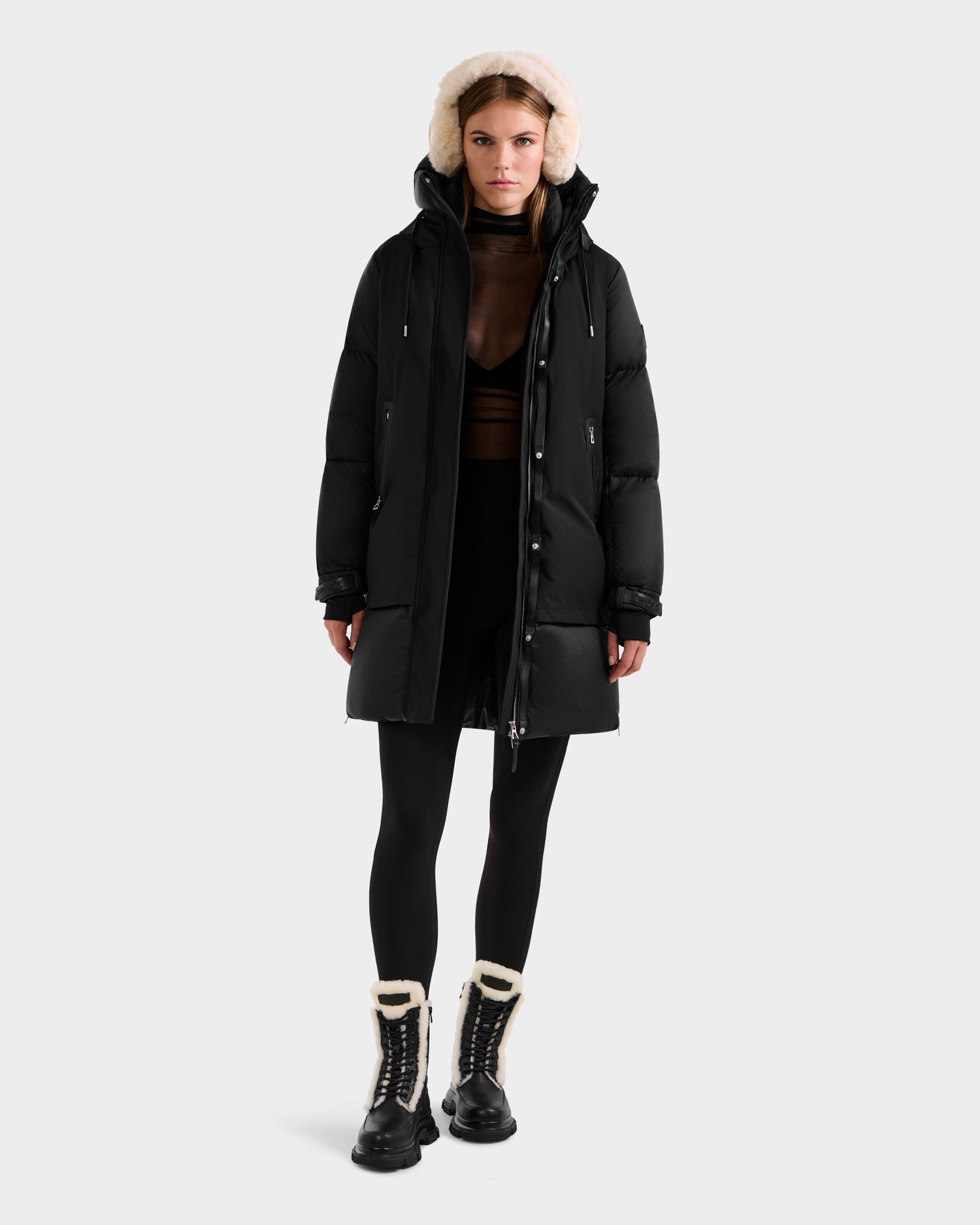 A woman faces forward, wearing the Rudsak LAYAN - 8124576 Black/Vanilla premium down puffer coat with a hood, black leggings, a sheer brown top, and black winter boots with white fur trim against a plain white background.