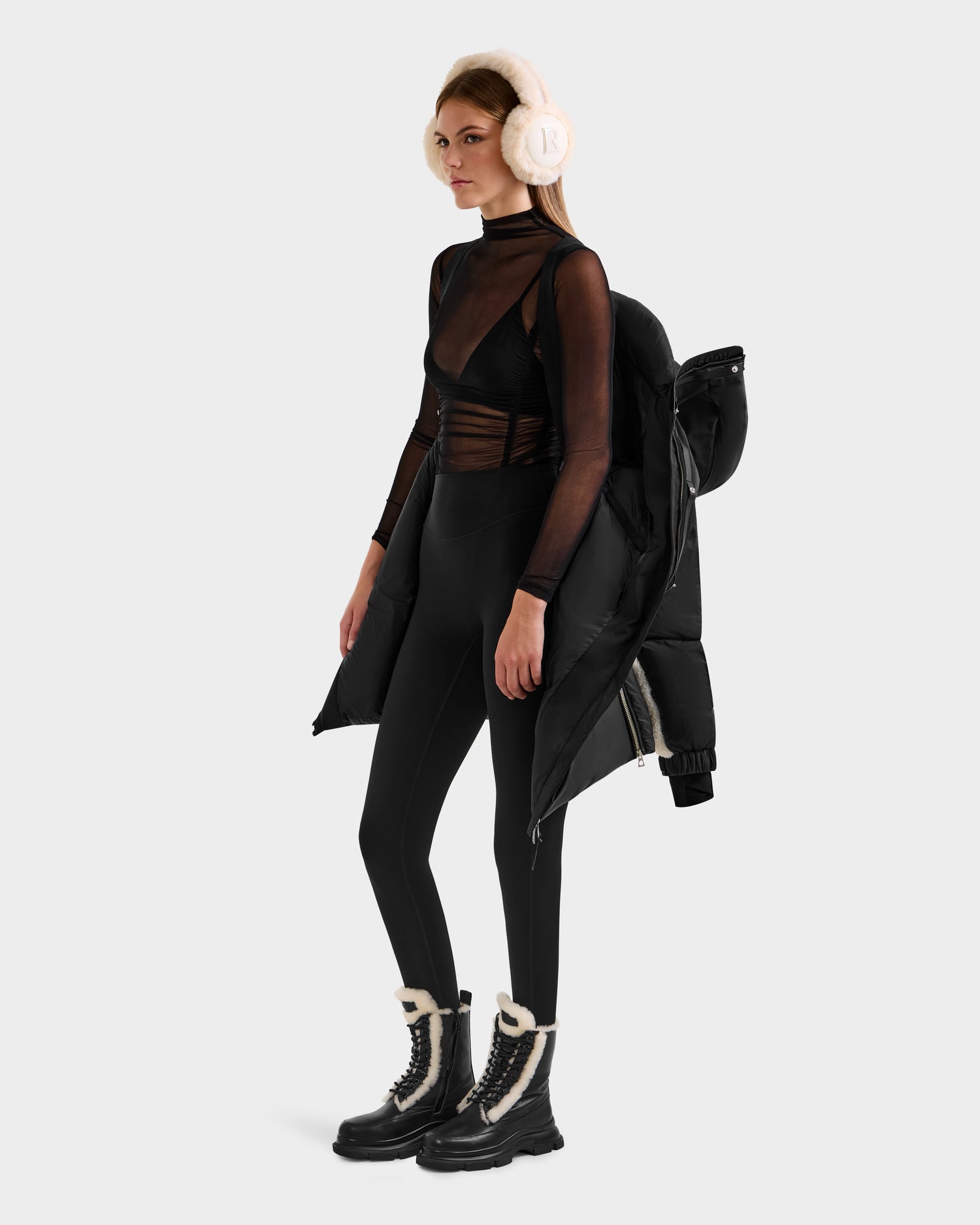 A woman in the Rudsak LAYAN - 8124576 Black/Vanilla premium down winter coat draped over one shoulder stands against a plain background, styled with black leggings, a sheer top, furry boots, and white earmuffs.
