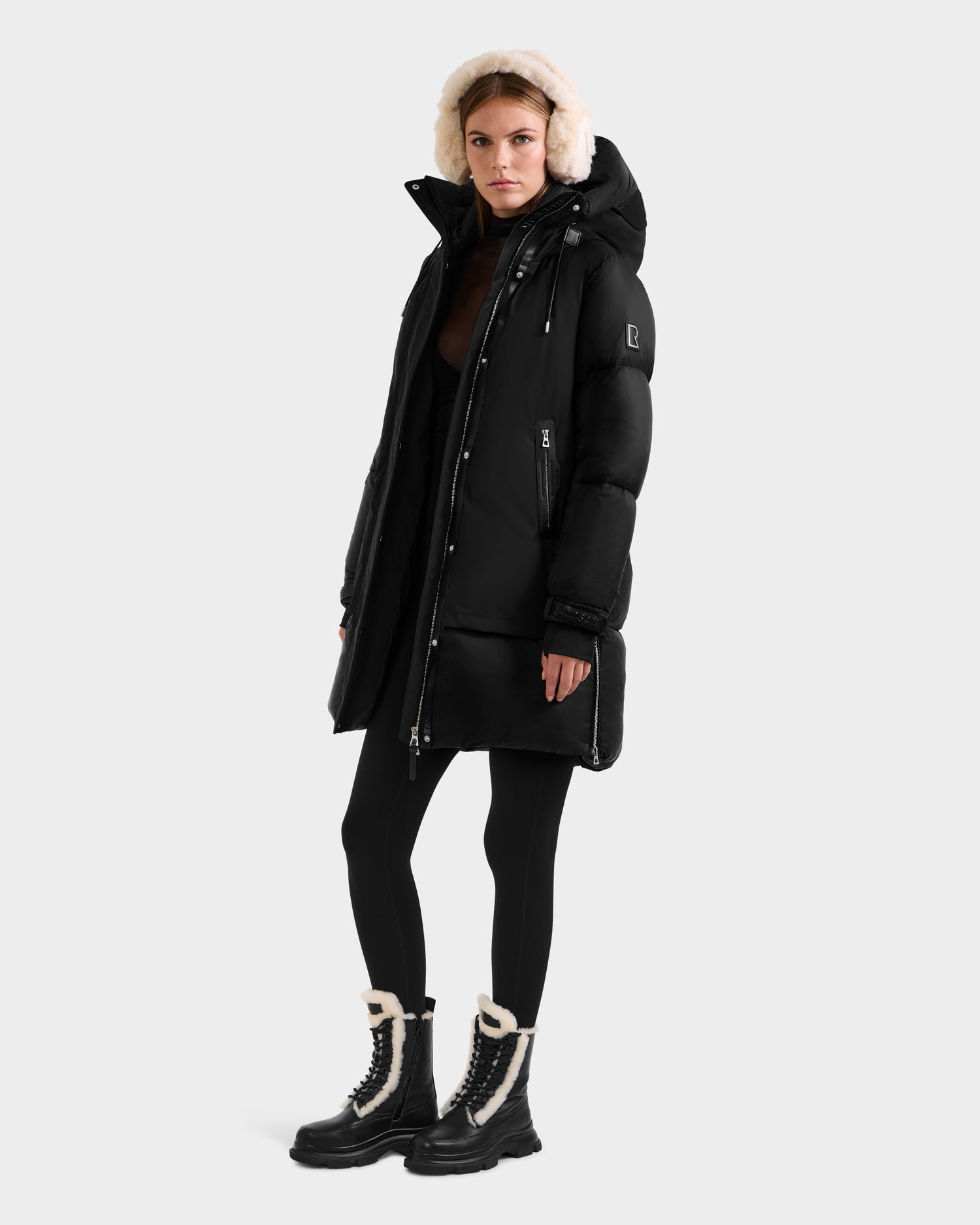 A woman in the Rudsak LAYAN - 8124576 Black/Vanilla down parka with a fur-lined hood stands against a plain background, pairing it with black leggings and black boots with white fur trim to showcase this stylish winter coat.