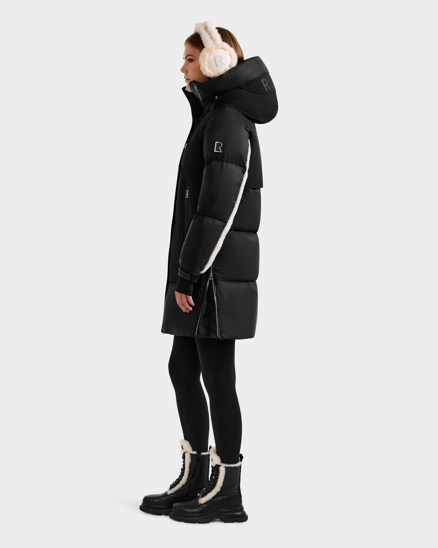 A woman models the Rudsak LAYAN - 8124576 Black/Vanilla premium down women’s winter coat, paired with black leggings, fur-lined boots, and beige earmuffs, standing in profile against a plain light background.