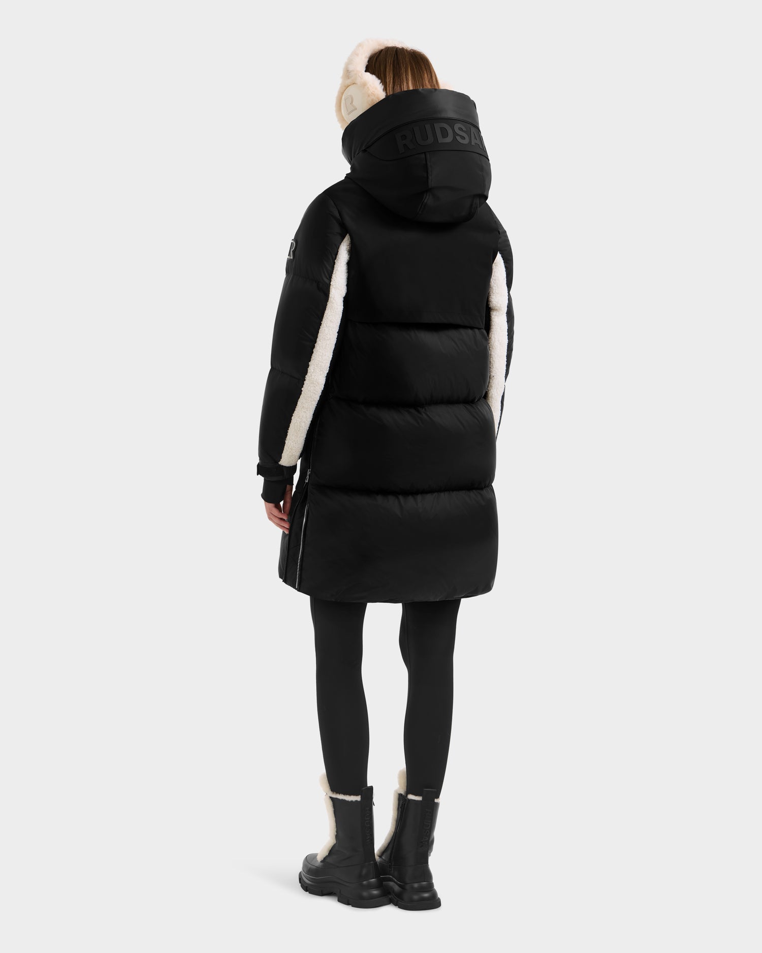 A person in the Rudsak LAYAN - 8124576 Black/Vanilla womens down parka with white shearling accents, black leggings, and black boots with white trim stands facing away against a plain light background—where style meets warmth.