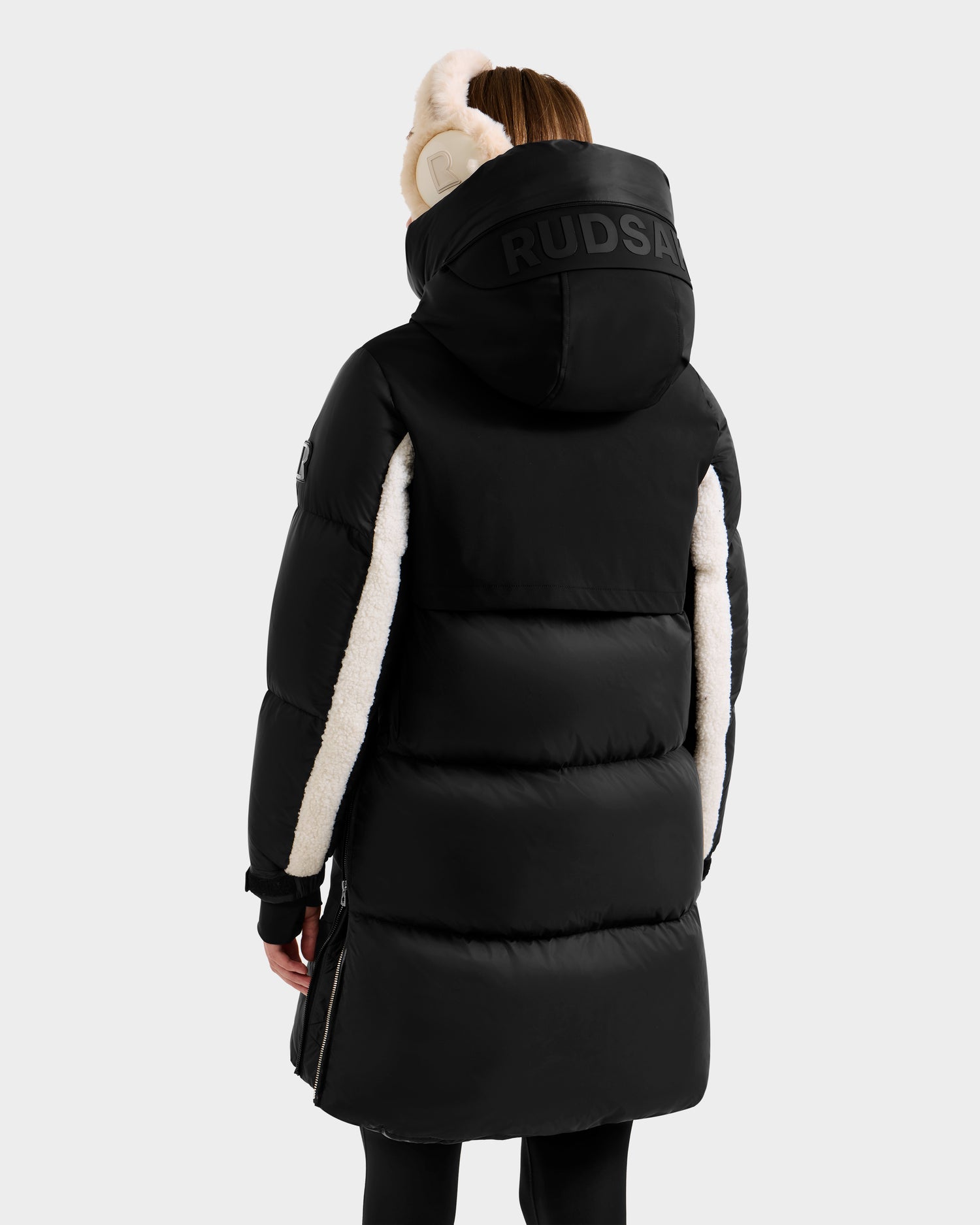 A person is seen from the back wearing the Rudsak LAYAN - 8124576 Black/Vanilla womens down parka, featuring a large hood with white fur lining and RUDSAK branding, set against a plain light-colored background.