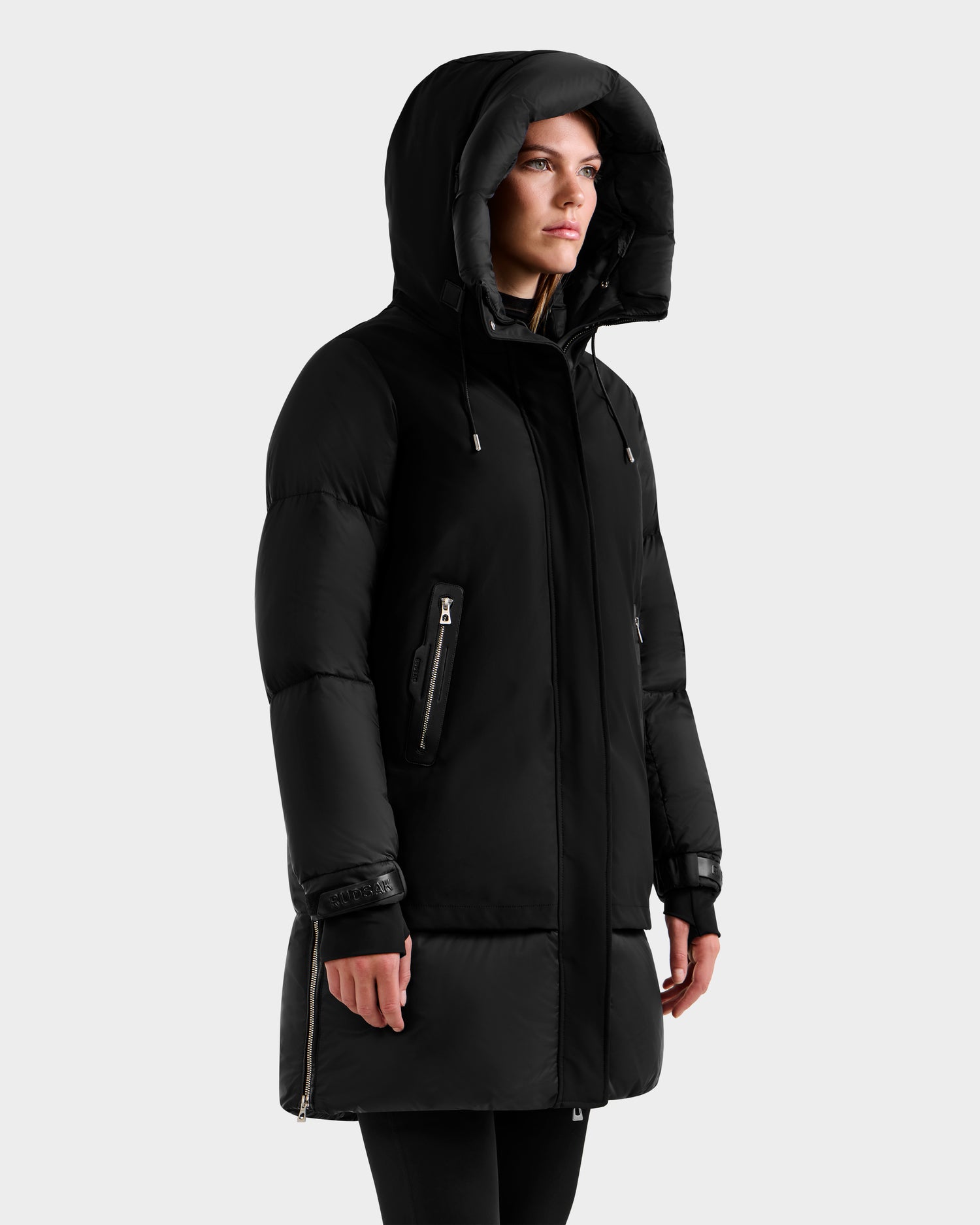 A person wearing the Rudsak LAYAN - 8124576 Black/Vanilla premium down long hooded puffer jacket with zippered pockets and a high collar stands against a plain light gray background.