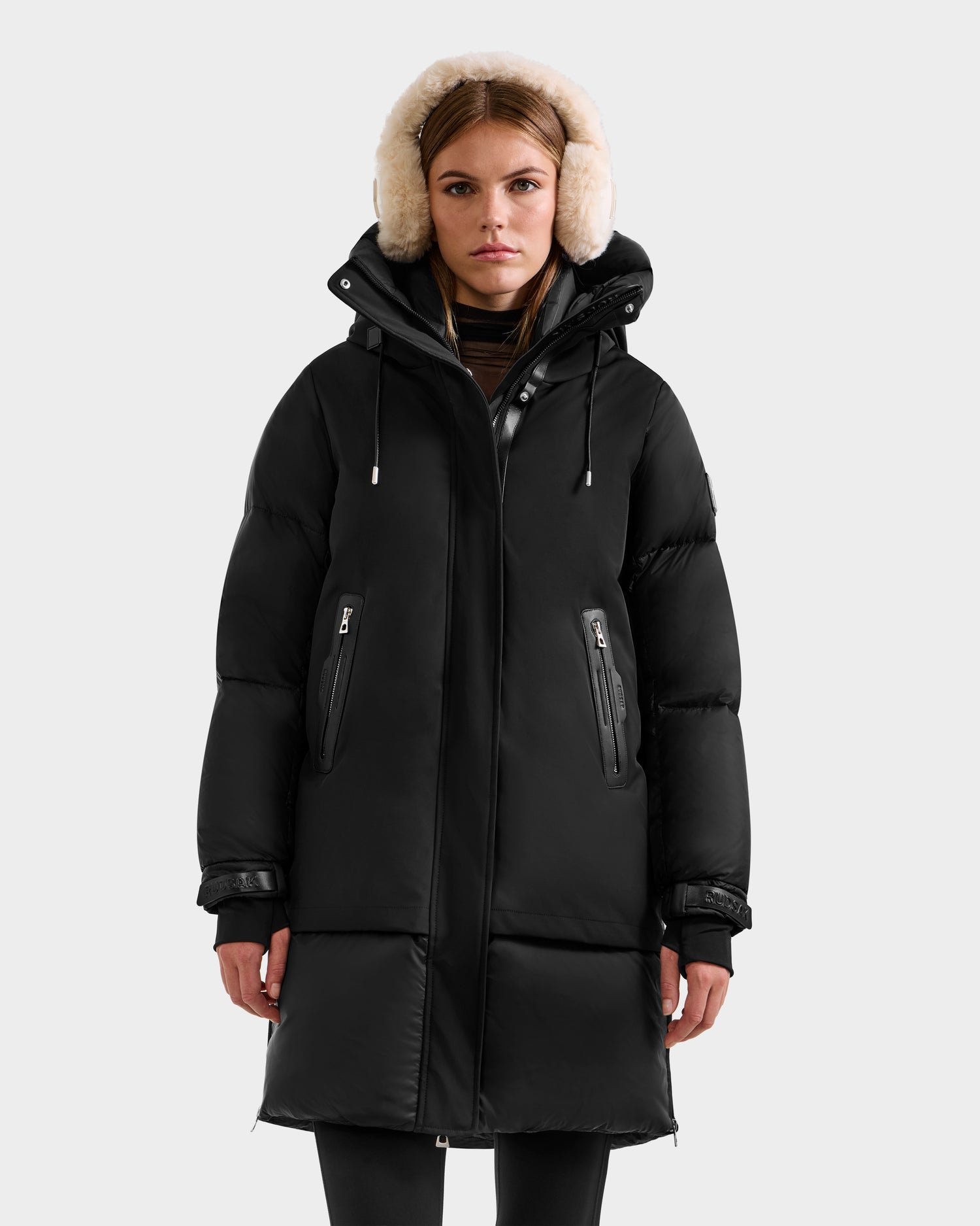 A woman wears the Rudsak LAYAN - 8124576 Black/Vanilla long down parka with a light-colored fur-lined hood, standing against a plain light gray background.