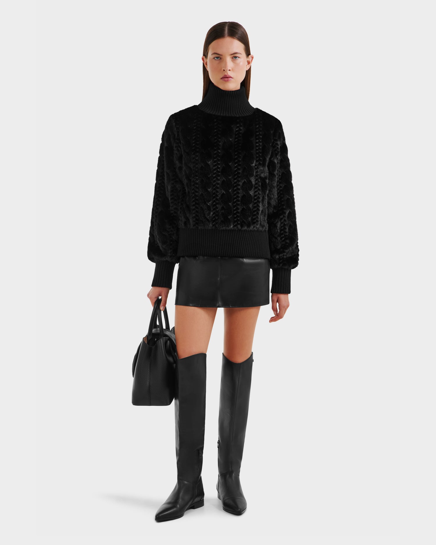 A woman with straight brown hair and a neutral expression wears the LAYLA F - 8525630 Black textured sweater by Rudsak, paired with a black leather mini skirt, knee-high boots, and holds a black handbag against a white background.