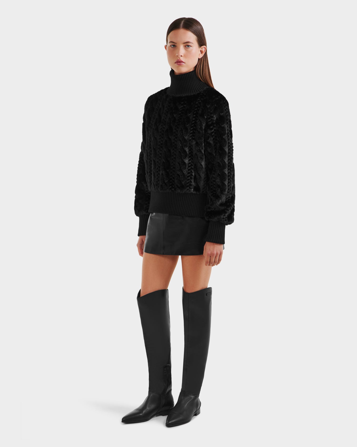 A woman with straight brown hair and a neutral expression wears the Rudsak LAYLA F - 8525630 Black après-ski style textured turtleneck sweater, paired with a black mini skirt and knee-high black boots, standing against a plain background.