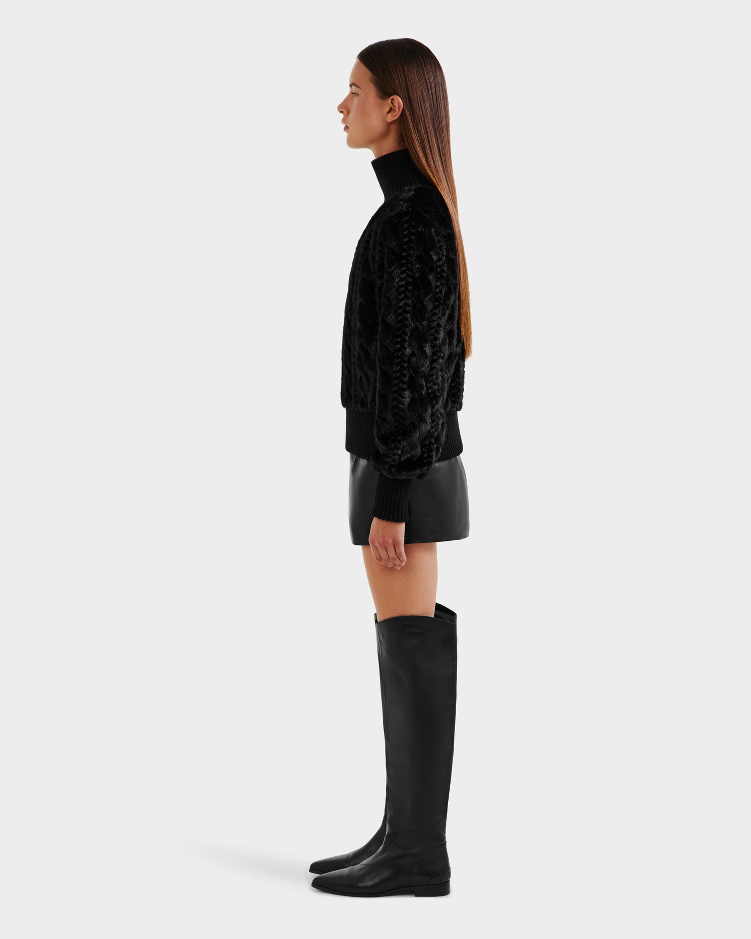 A woman with straight, loose hair is seen in profile against a plain background, wearing the Rudsak LAYLA F - 8525630 Black cable knit-inspired turtleneck sweater, paired with a black leather mini skirt and knee-high black boots.