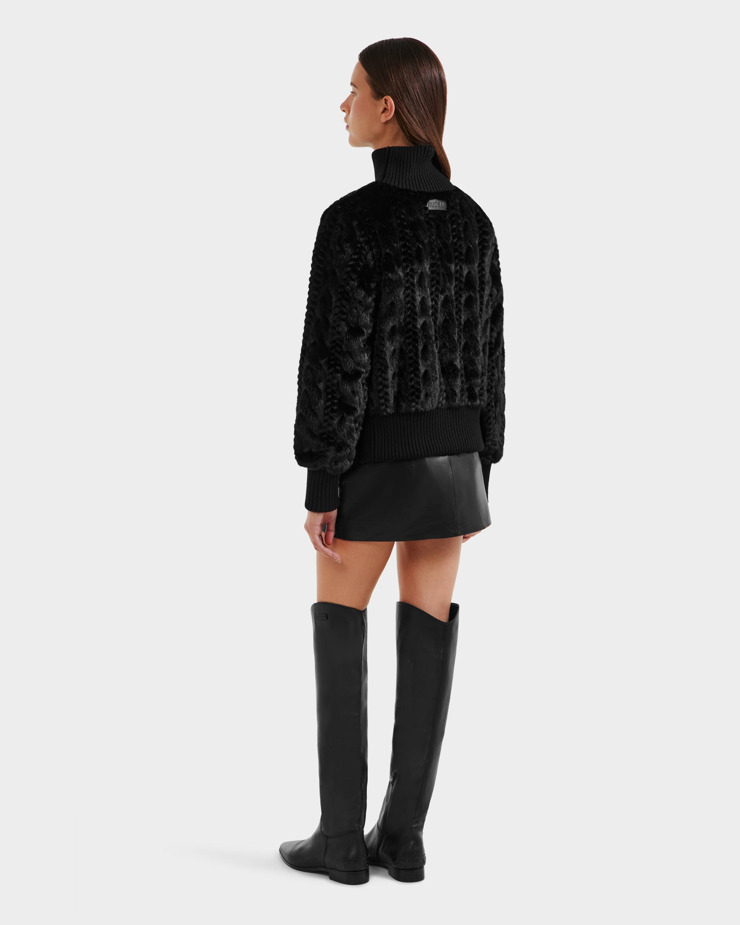 A woman with straight brown hair, hands by her sides, models the Rudsak LAYLA F - 8525630 Black textured jacket with a black mini skirt and knee-high boots against a plain light background.