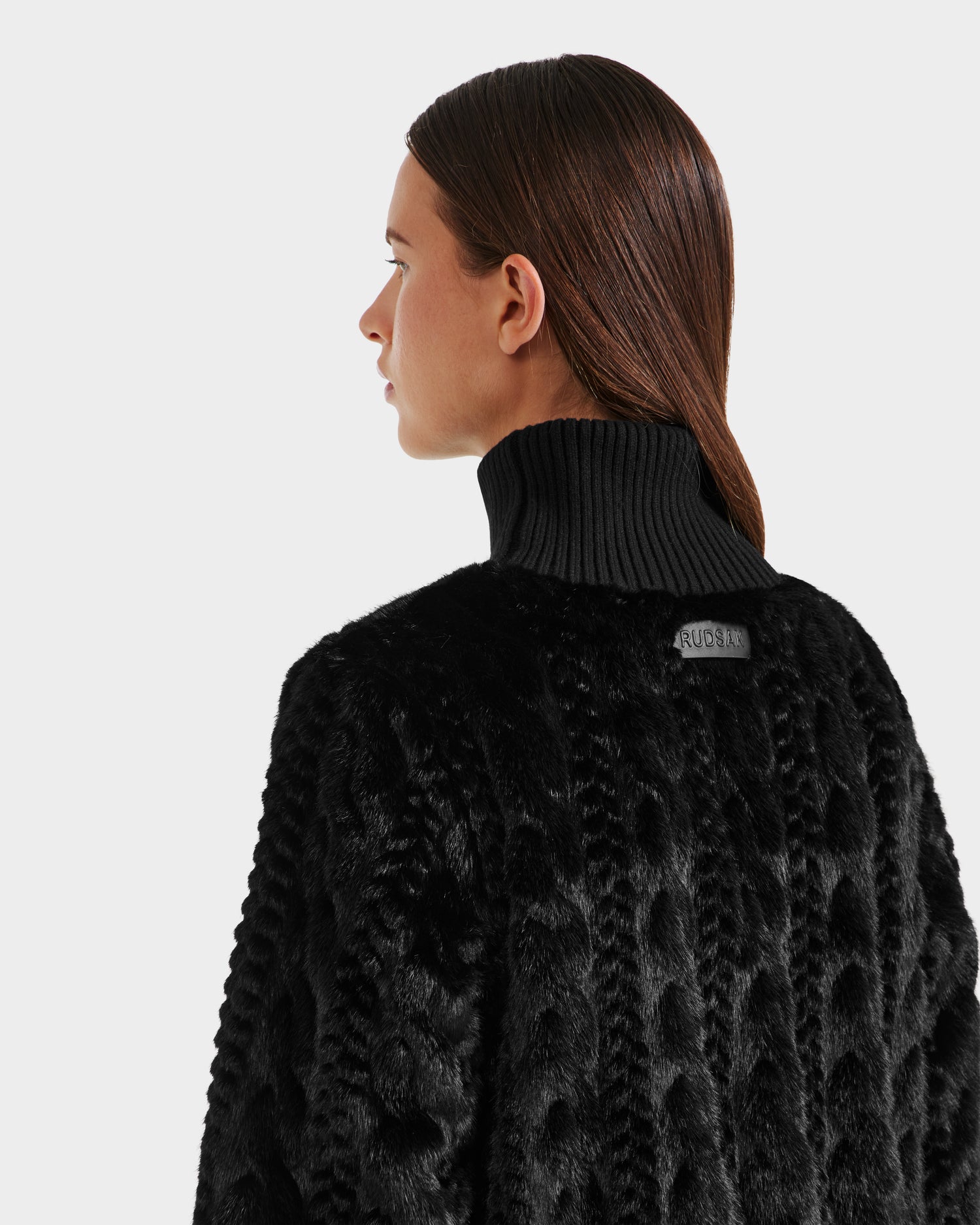 A person with straight brown hair models the Rudsak LAYLA F - 8525630 Black women’s faux fur turtleneck coat with black ribbed collar, shown from the back and side against a plain light background, evoking après-ski style.