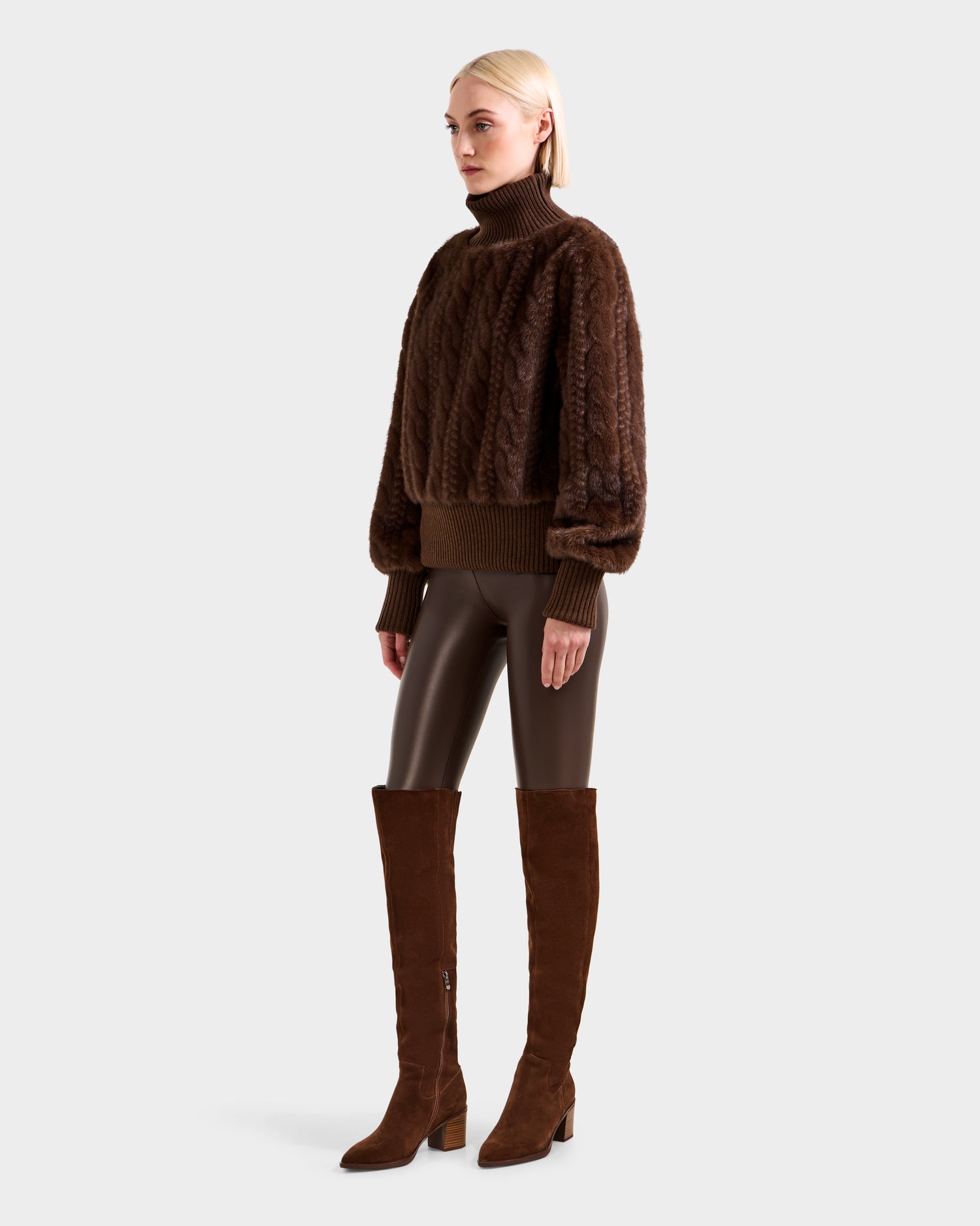 A blonde woman wears the Rudsak LAYLA F - 8525630 Mocha women’s faux fur turtleneck sweater with dark brown leather pants and knee-high brown suede boots, standing against a plain light gray background.