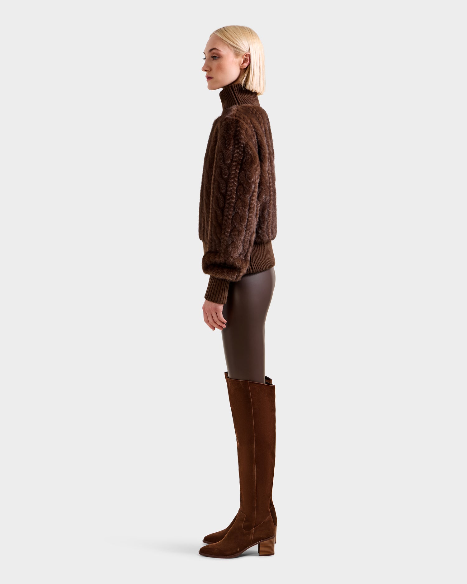 A woman with blonde hair stands in profile against a light gray background, wearing the Rudsak LAYLA F - 8525630 Mocha chunky cable-knit turtleneck sweater styled with brown leggings and knee-high brown suede boots.