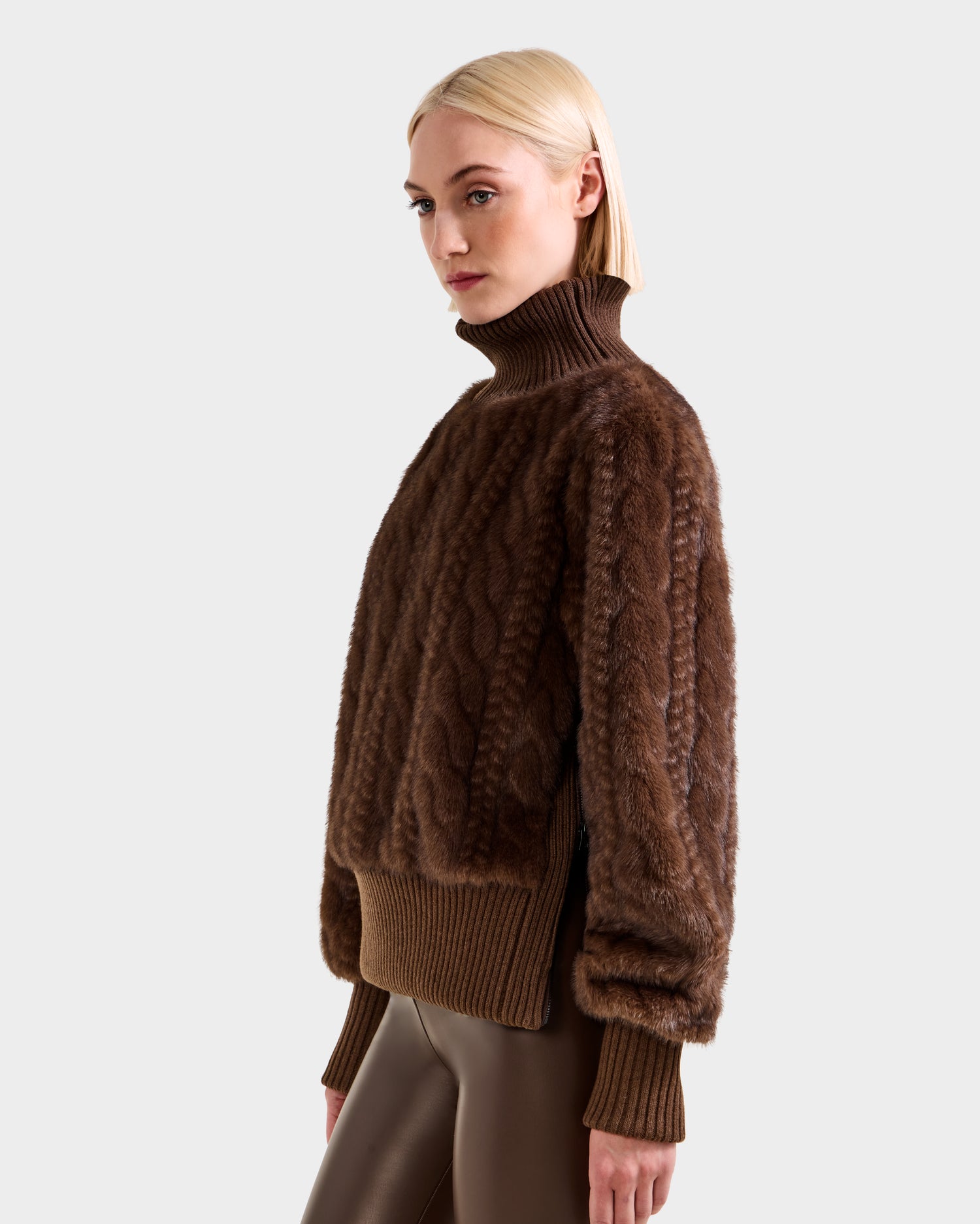 A woman with blonde hair wears the Rudsak LAYLA F - 8525630 Mocha après-ski turtleneck sweater with ribbed cuffs and hem, paired with shiny brown leggings. She stands against a plain light gray background, gazing slightly to the side.