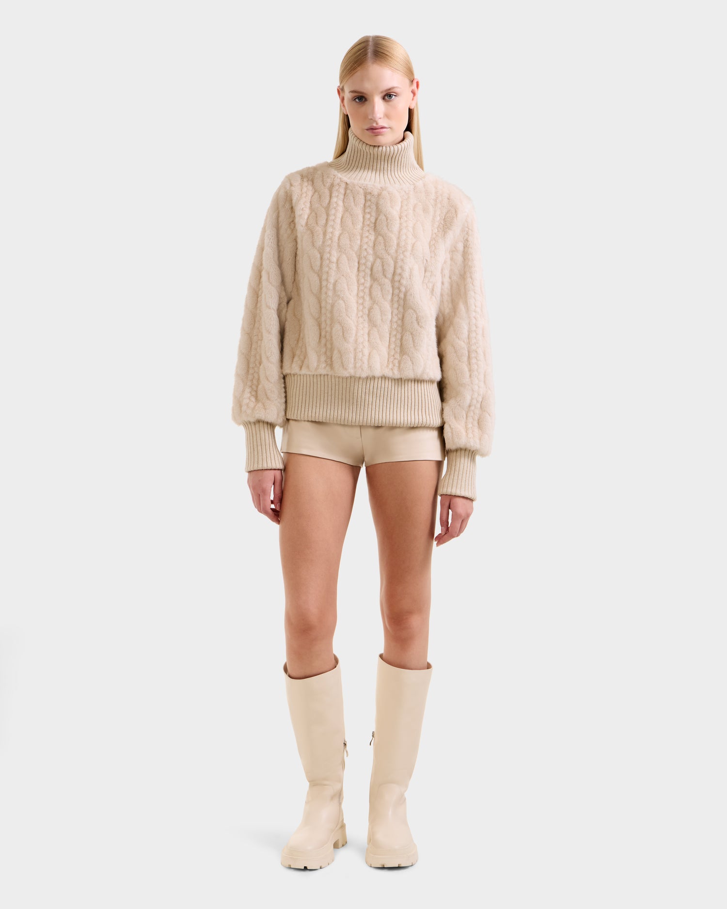 A woman faces forward against a white background, wearing the Rudsak LAYLA F - 8525630 Vanilla cable knit-inspired faux fur turtleneck sweater with beige shorts and cream knee-high boots for an effortless après-ski look.
