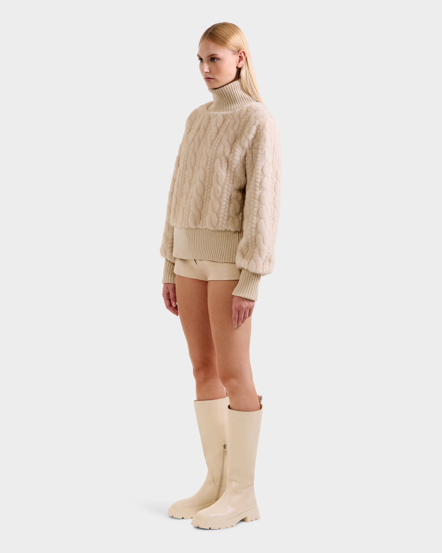 A woman with straight blonde hair and a neutral expression stands against a plain background, wearing Rudsak's LAYLA F - 8525630 Vanilla faux fur cream sweater, beige shorts, and tall beige rain boots. She looks slightly to the side.