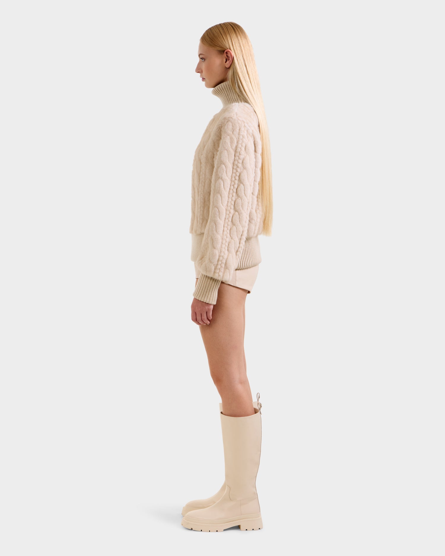 A woman with long blonde hair stands in profile, embracing après-ski style in Rudsak’s LAYLA F - 8525630 Vanilla: a beige cable-knit sweater, matching shorts, and knee-high cream boots against a plain light background.