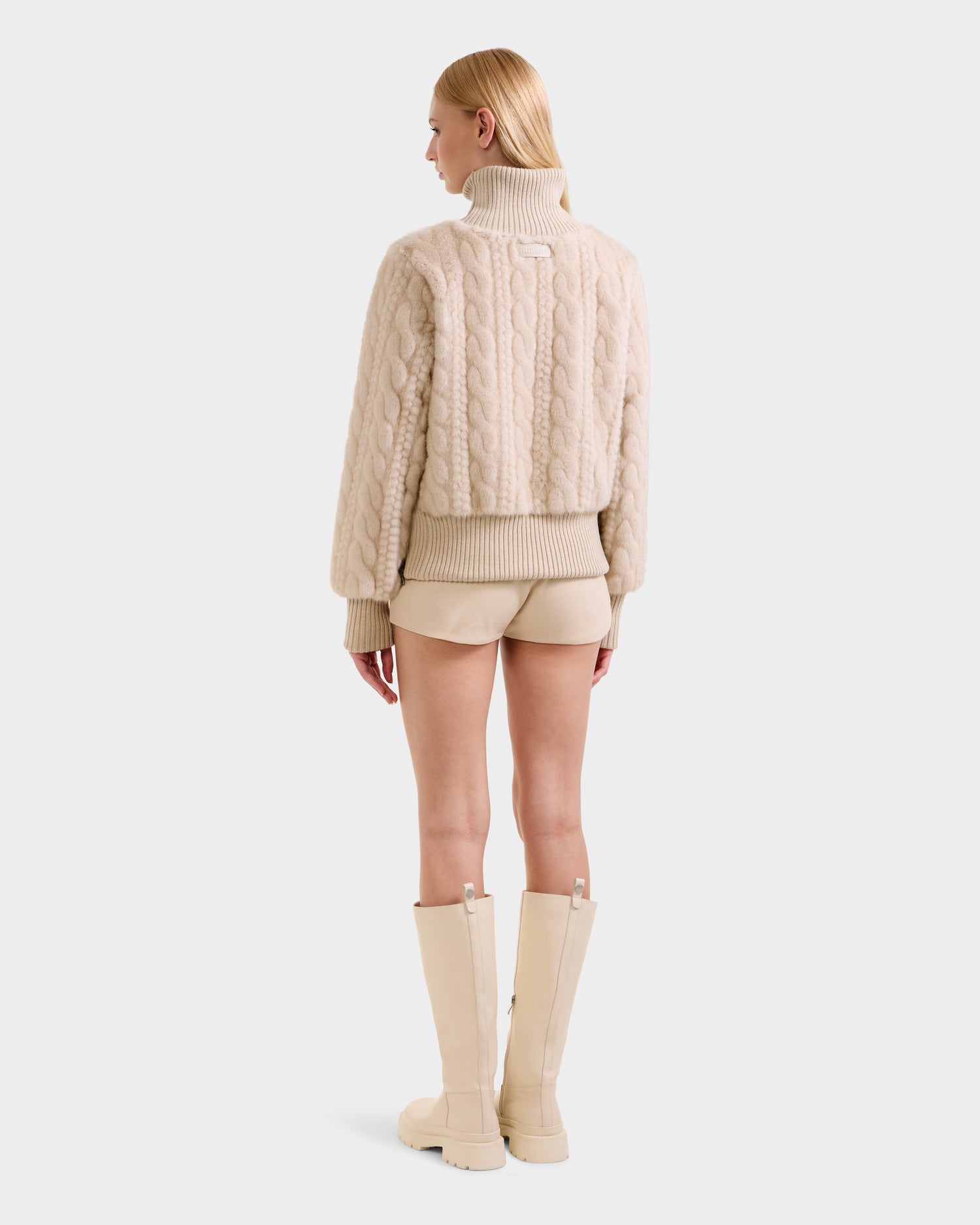 A woman with straight blonde hair stands facing away, wearing the Rudsak LAYLA F - 8525630 Vanilla beige cable-knit turtleneck sweater, beige shorts, and knee-high cream boots against a plain white background.