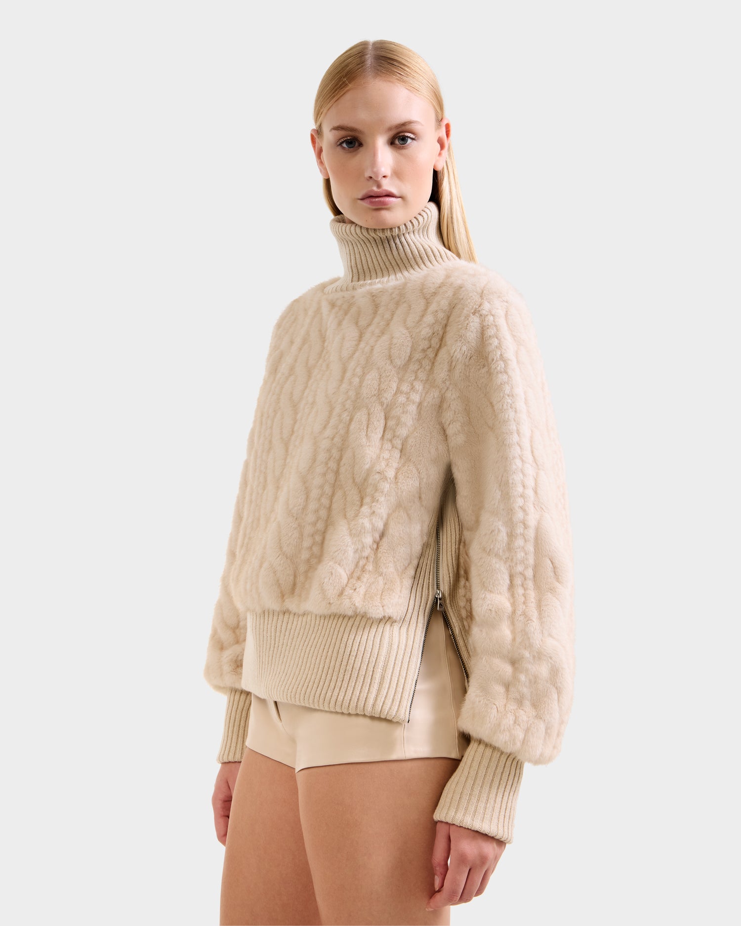 A woman with straight blonde hair wears the Rudsak LAYLA F - 8525630 Vanilla cable-textured après-ski sweater with ribbed cuffs and hem, paired with beige shorts, standing against a plain light background.