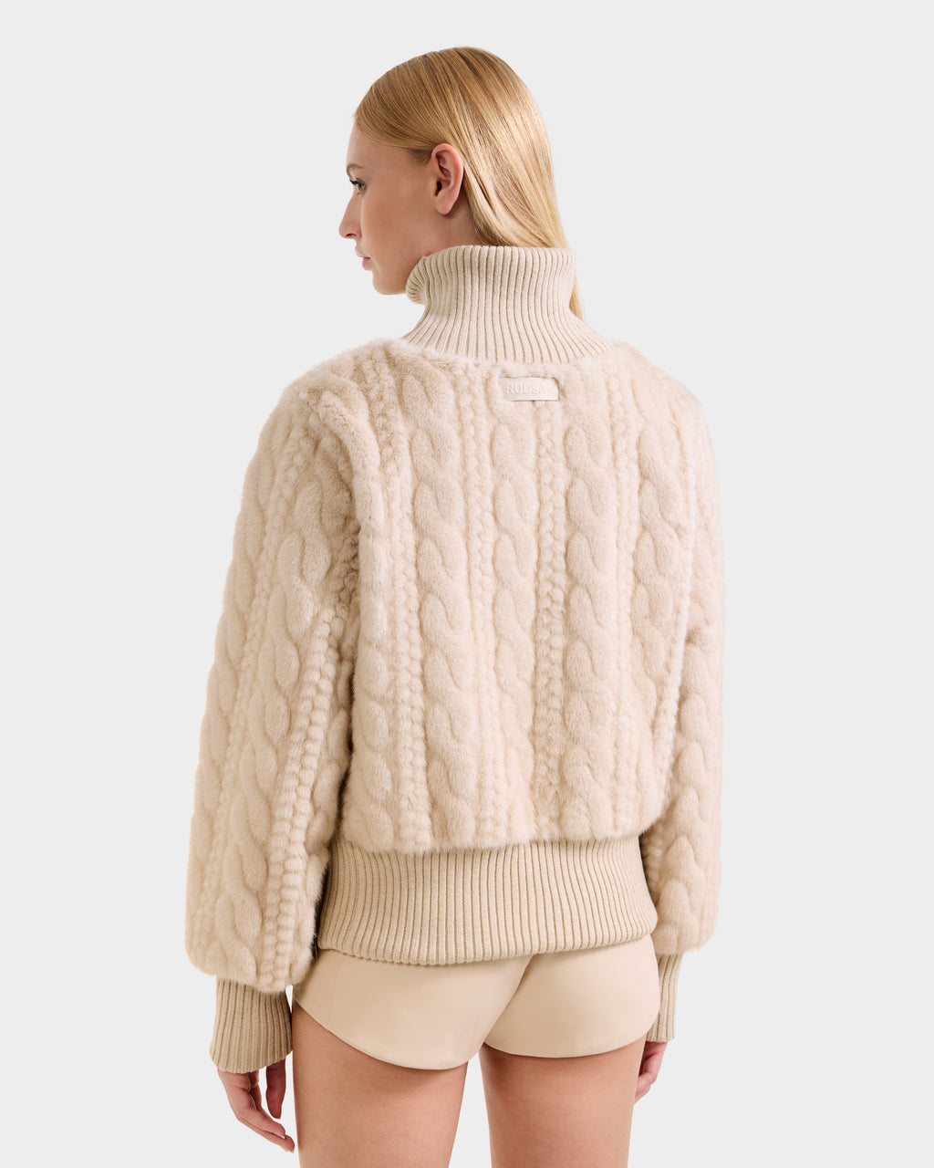 A woman with straight blonde hair models the Rudsak LAYLA F - 8525630 Vanilla cable knit turtleneck sweater, styled après-ski, paired with beige shorts and shown from behind against a plain light background.