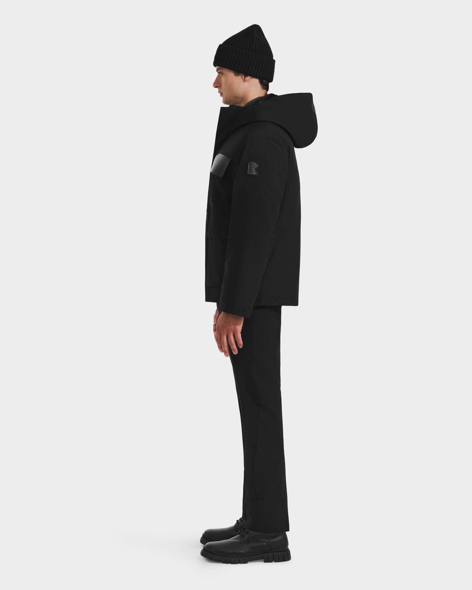 A person stands in profile facing left, wearing the Rudsak LAZIO - 6125527 Black winter jacket, paired with black pants and shoes, against a plain white background.