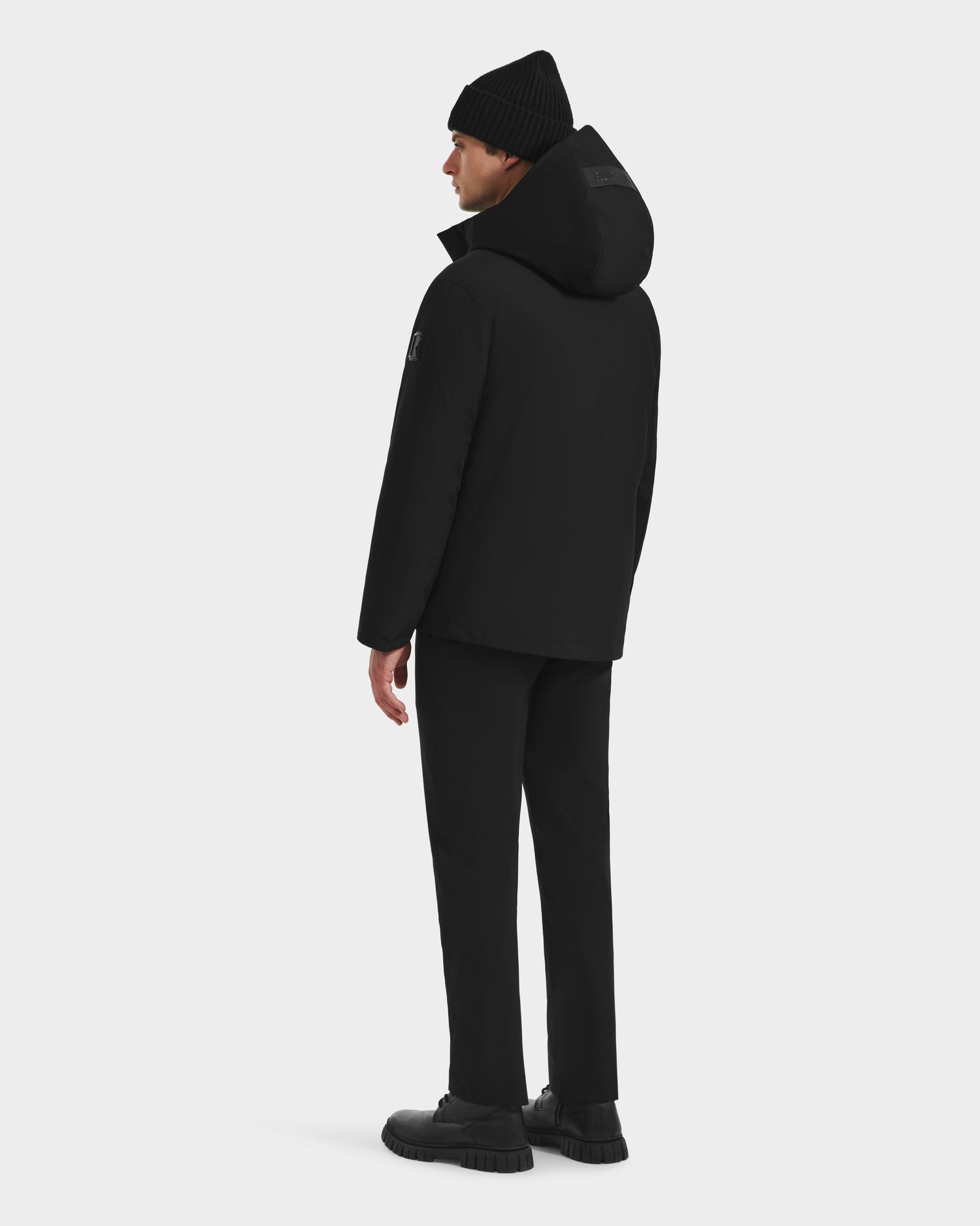A man faces away wearing the Rudsak LAZIO - 6125527 Black winter jacket, paired with black pants, shoes, and a beanie—delivering sleek cold-weather protection against a plain white background.