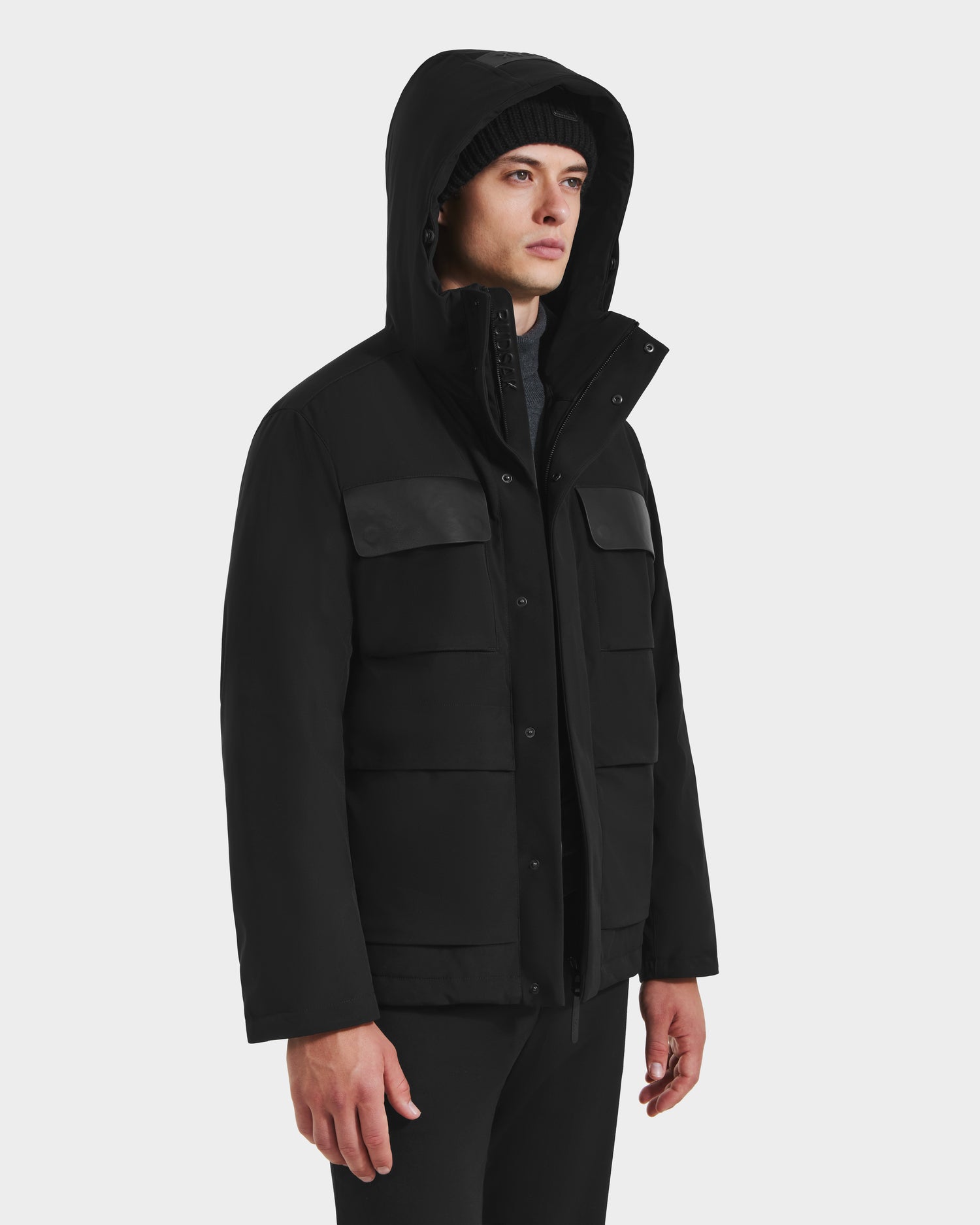 A person wears the Rudsak LAZIO - 6125527 Black men’s parka with multiple front pockets, standing against a plain white background and looking slightly to the side, showcasing cold-weather style.