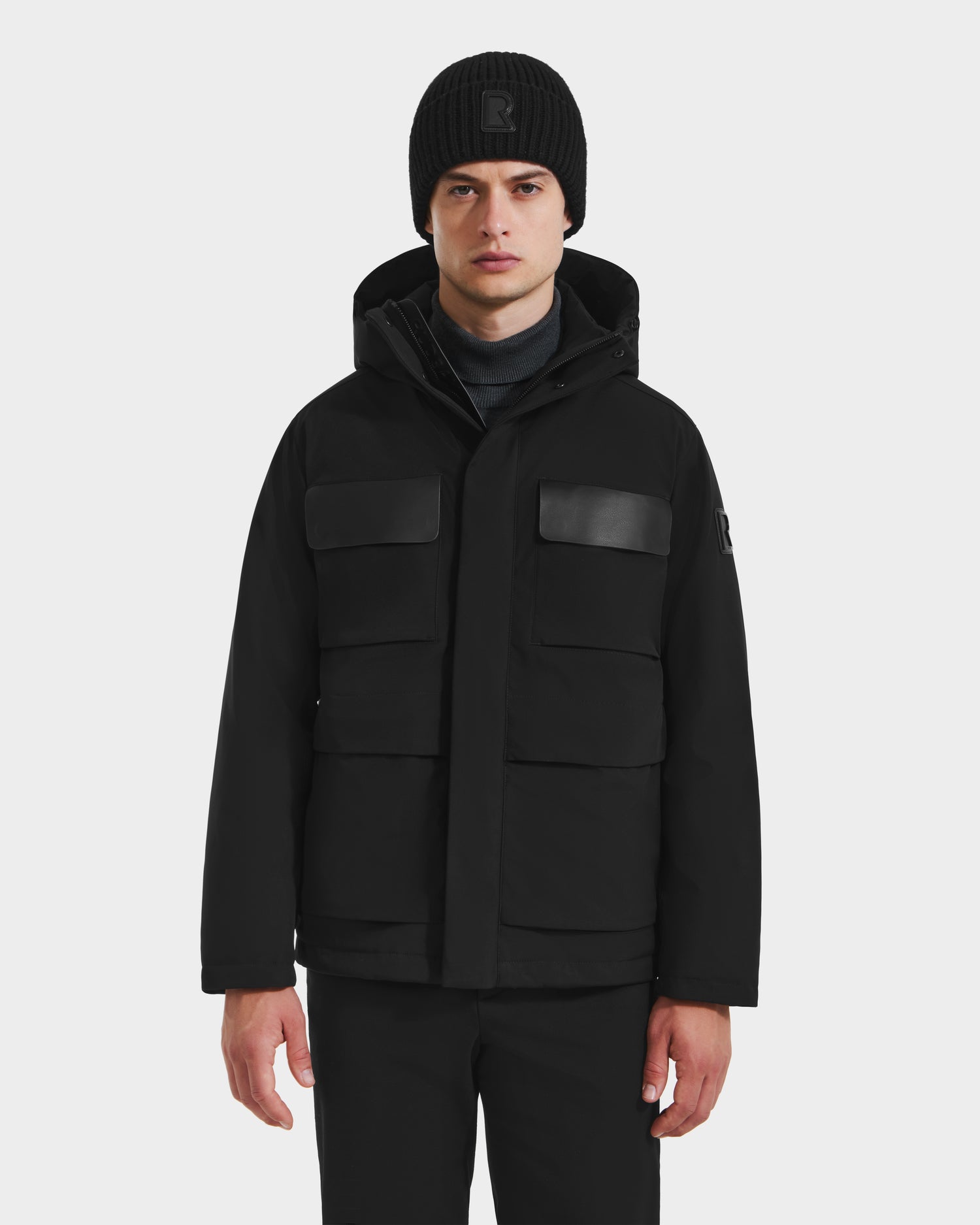 A man wearing the Rudsak LAZIO - 6125527 Black parka with large front pockets and a black beanie stands facing forward against a white background, highlighting stylish cold-weather protection.