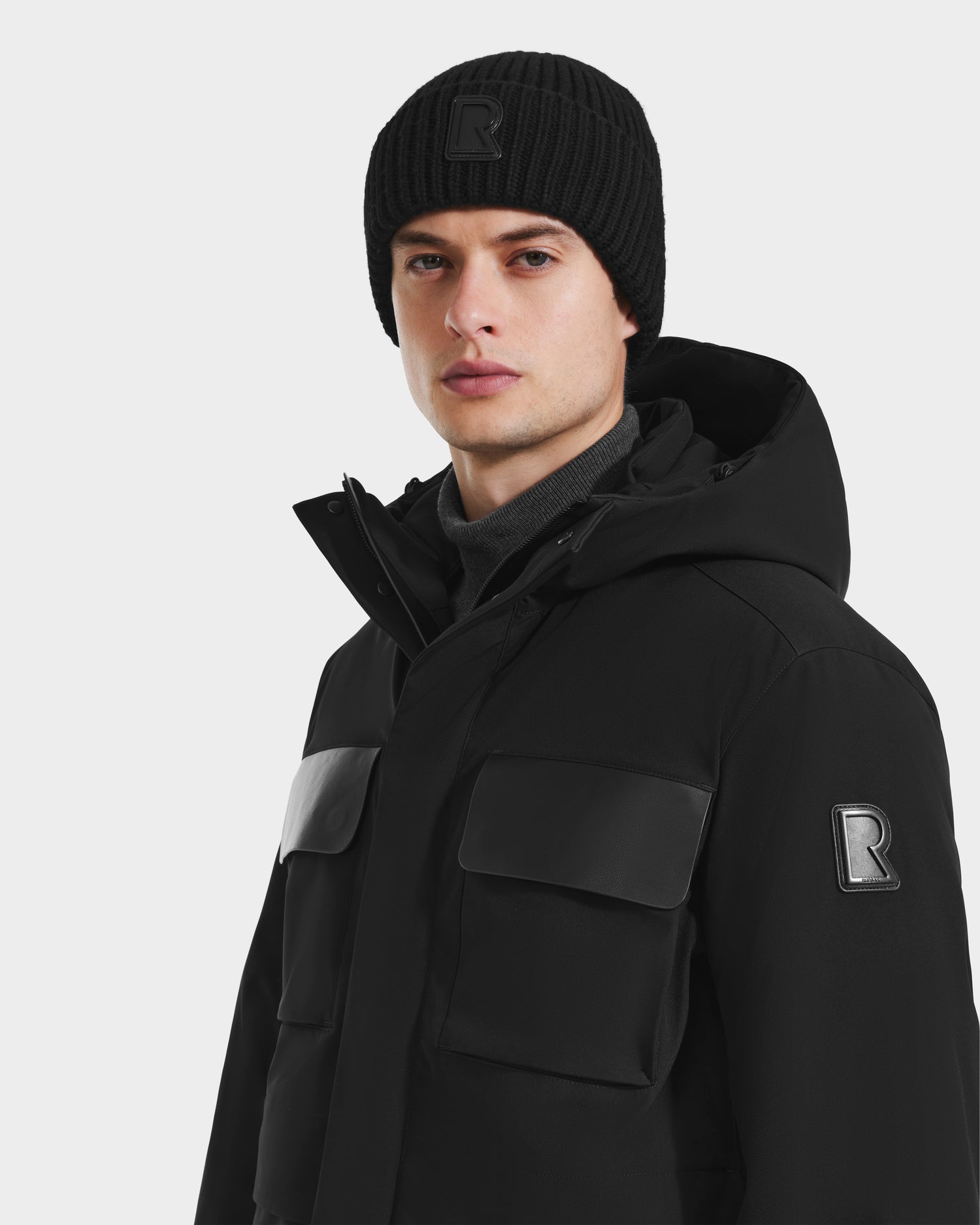 A person in the Rudsak LAZIO - 6125527 Black hooded winter jacket with large front pockets and a black knit beanie, both with the prominent R logo, stands against a white background—stylish and warm for cold weather.