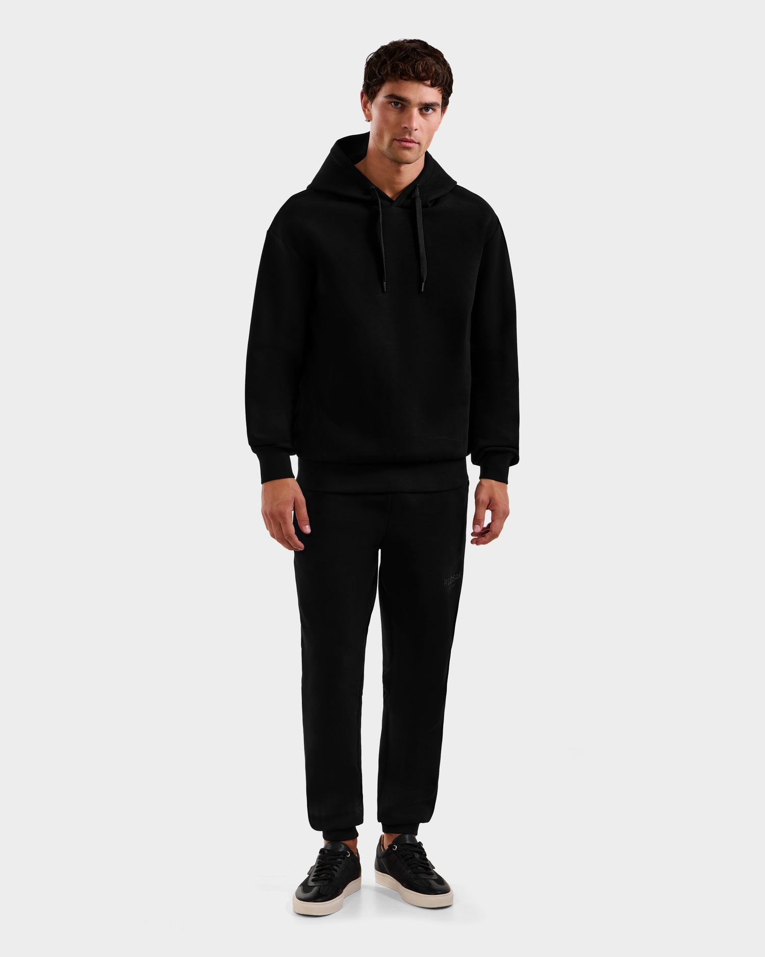 A young man stands facing forward, wearing the Rudsak LEROY - U525509 Black M hoodie with black jogger pants and black sneakers with white soles, against a plain light gray background.
