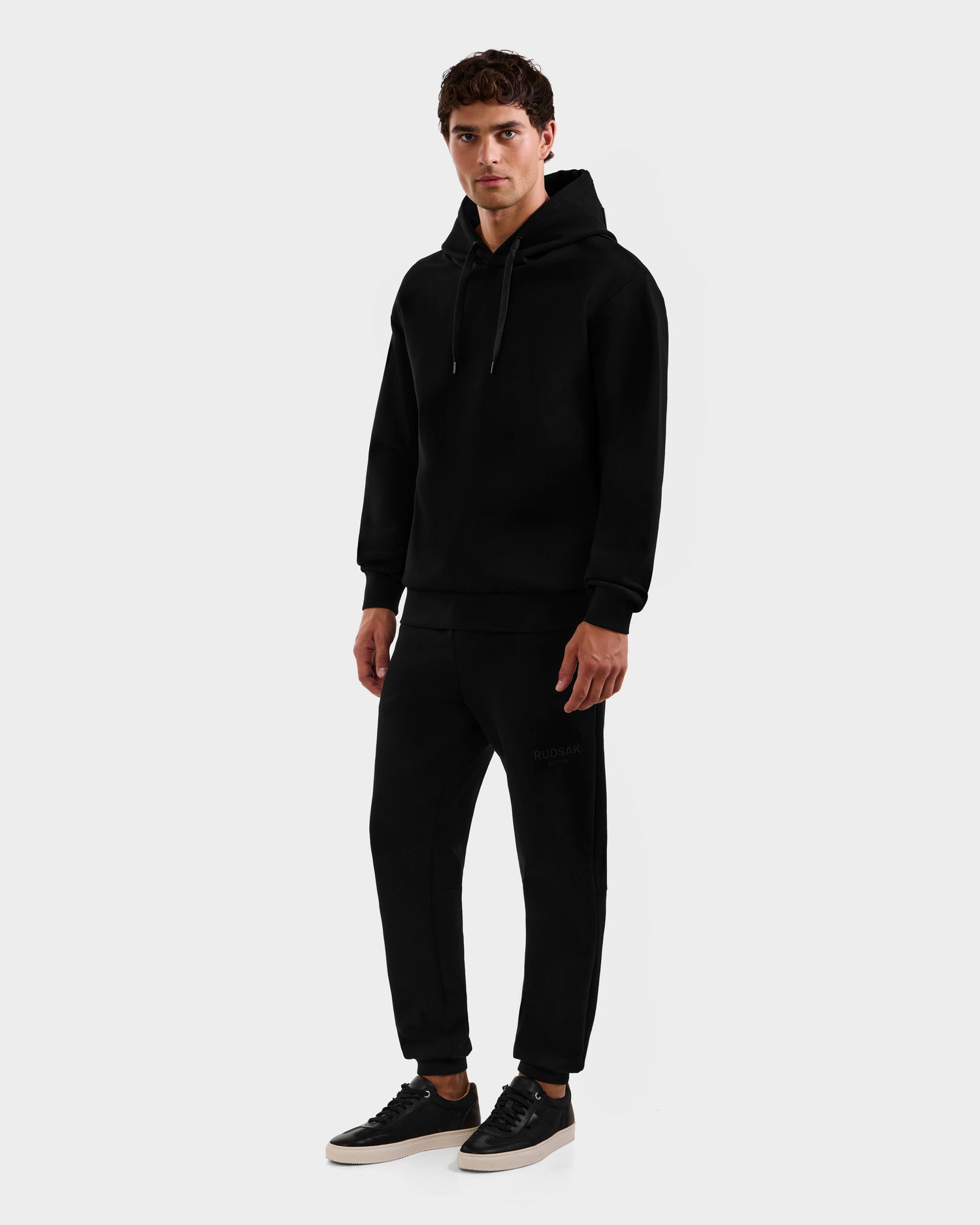 A person faces forward against a light gray background, wearing the Rudsak LEROY - U525509 Black M unisex hoodie, paired with black jogger pants and black sneakers with white soles.