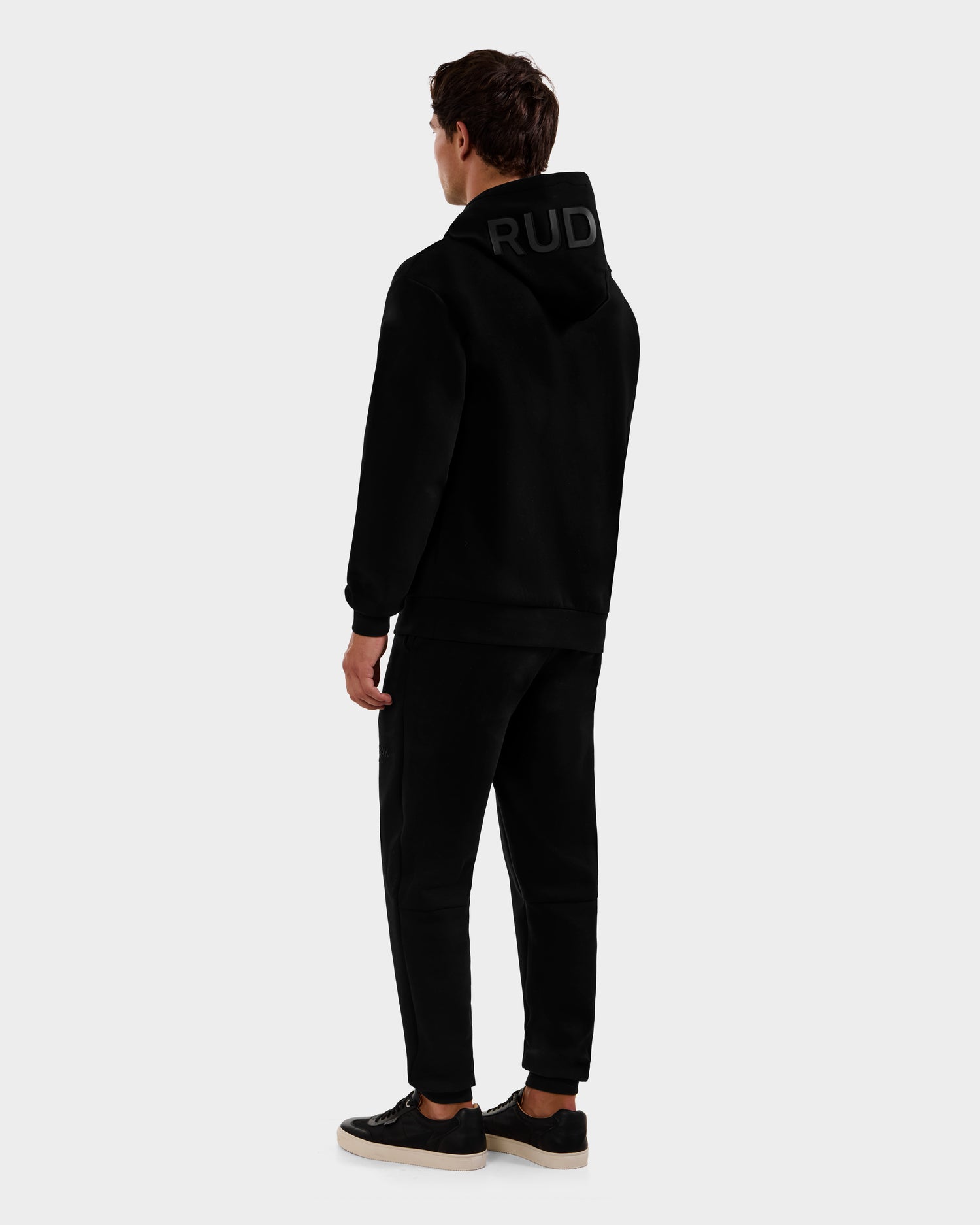 A person faces away, wearing the Rudsak LEROY - U525509 Black M hoodie with "RUD" on the hood, paired with black pants and black sneakers with white soles, set against a light gray background.