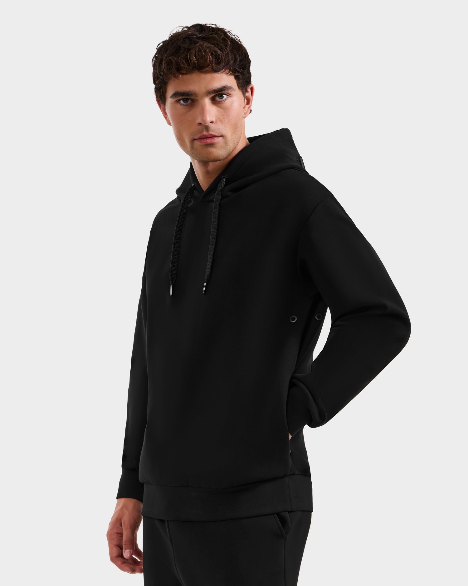 A young man with short curly hair models the Rudsak LEROY - U525509 Black M unisex hoodie, featuring black drawstrings and a front pocket. He stands against a light gray background, gazing slightly left with one hand in his pocket.