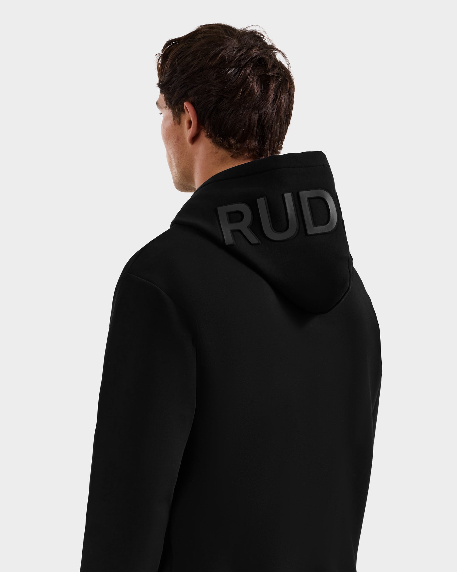 A person faces away, wearing the Rudsak LEROY - U525509 Black M premium hoodie, featuring bold black "RUD" lettering on the hood. A plain, light background highlights this stylish, gender-inclusive piece.