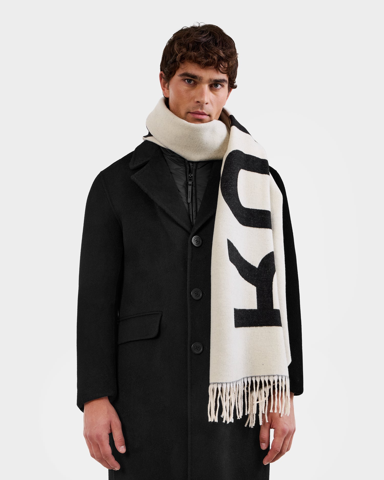 A man wears the LEV - U425506 Black coat by Rudsak and a cream-colored logo scarf with black lettering and fringe, an ideal cold-weather accessory, standing against a plain white background.