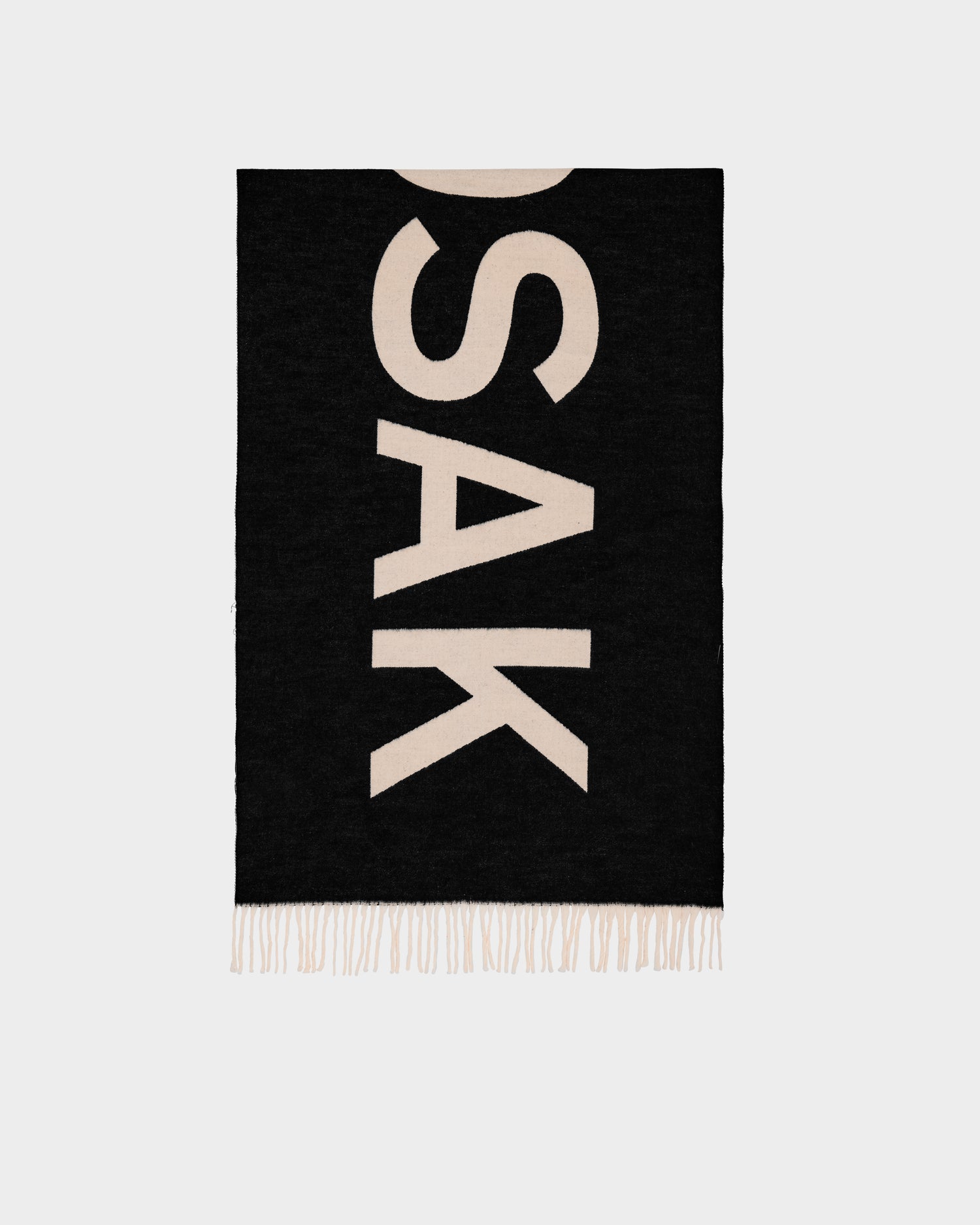 The LEV - U425506 Black by Rudsak is a reversible unisex scarf in black, featuring bold white SAK lettering printed vertically and white fringe at the ends. Background is plain and light-colored.