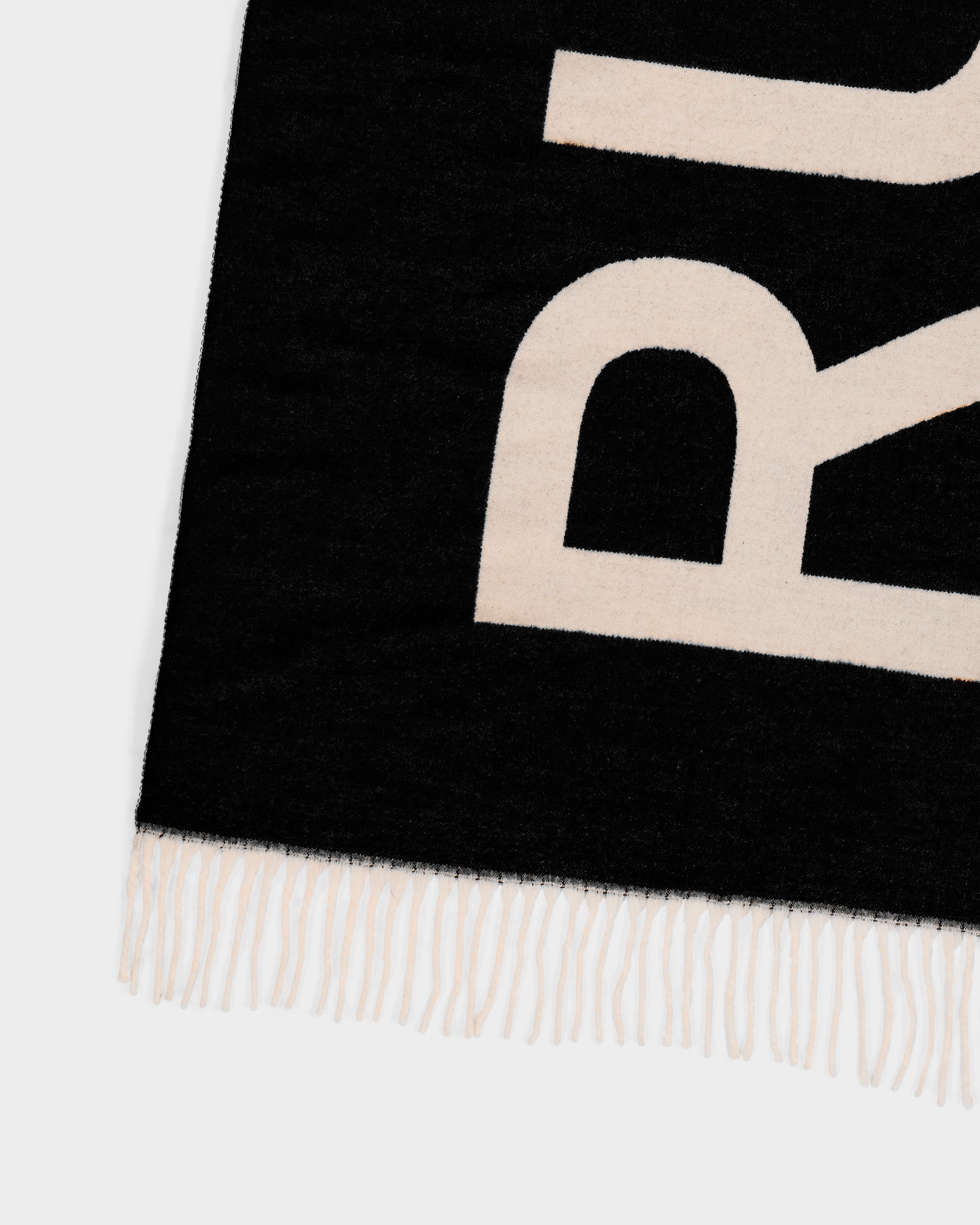 The Rudsak LEV - U425506 Black is a black woven reversible unisex scarf featuring cream abstract lettering, fringed edges, and a decorative border. Only part of this cold-weather accessory is shown, with partial letters visible.