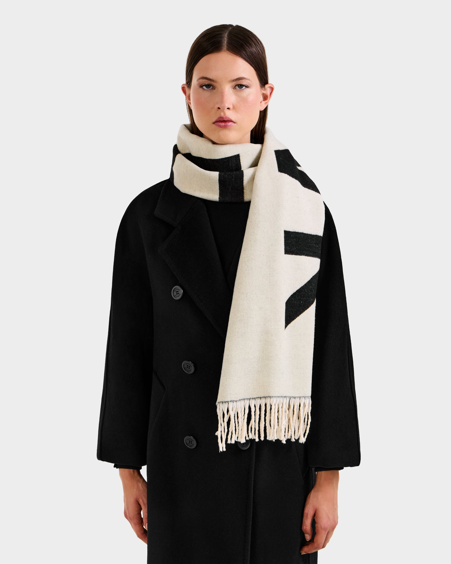 A woman in a long black coat models the Rudsak LEV - U425506 Black—a reversible unisex scarf featuring bold black patterns, cream tones, fringe ends, and logo details—against a plain light background.
