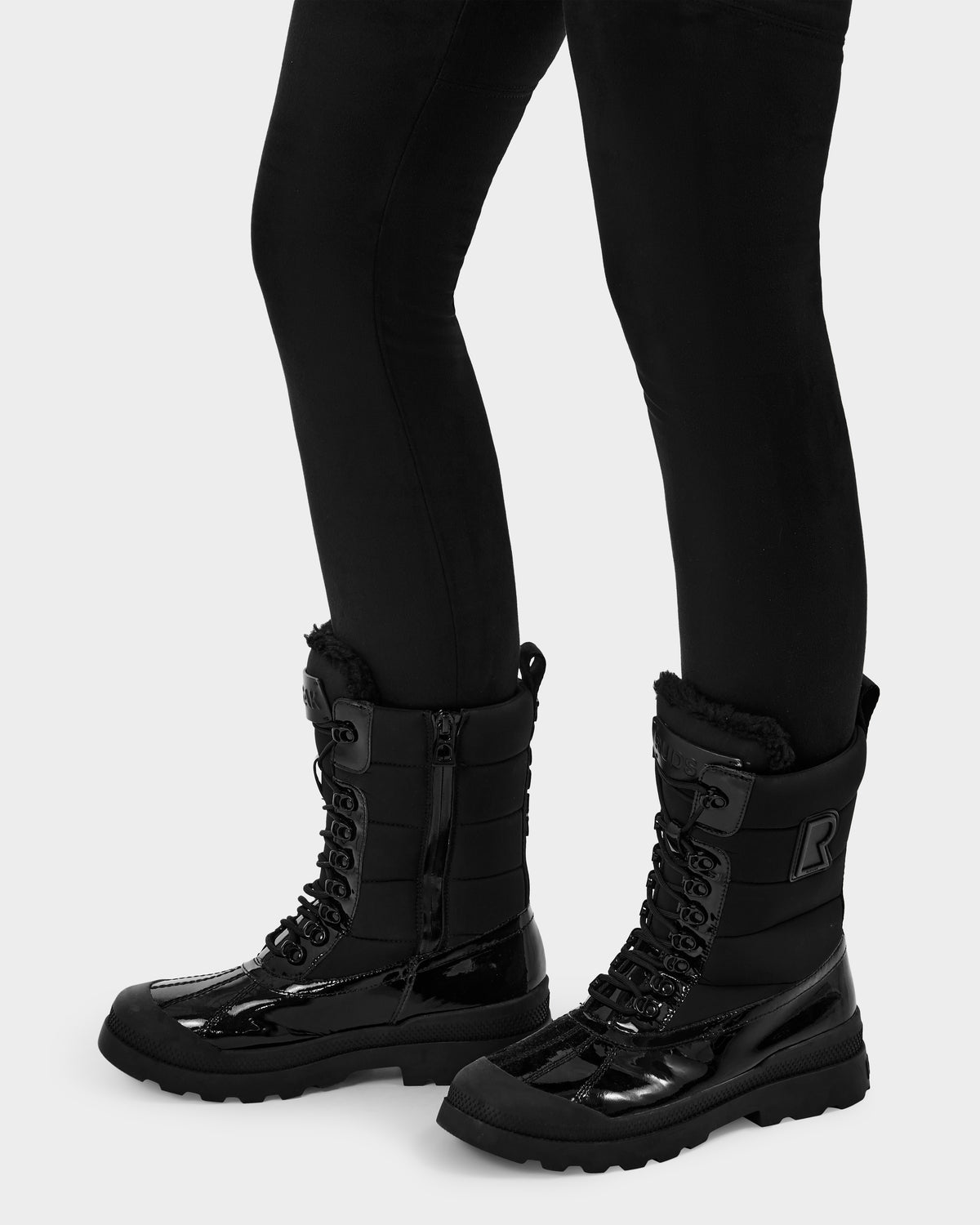 A person wearing black leggings and Rudsak LEX - 8224534 Black women’s winter boots with thick soles and fur lining stands against a plain light background.