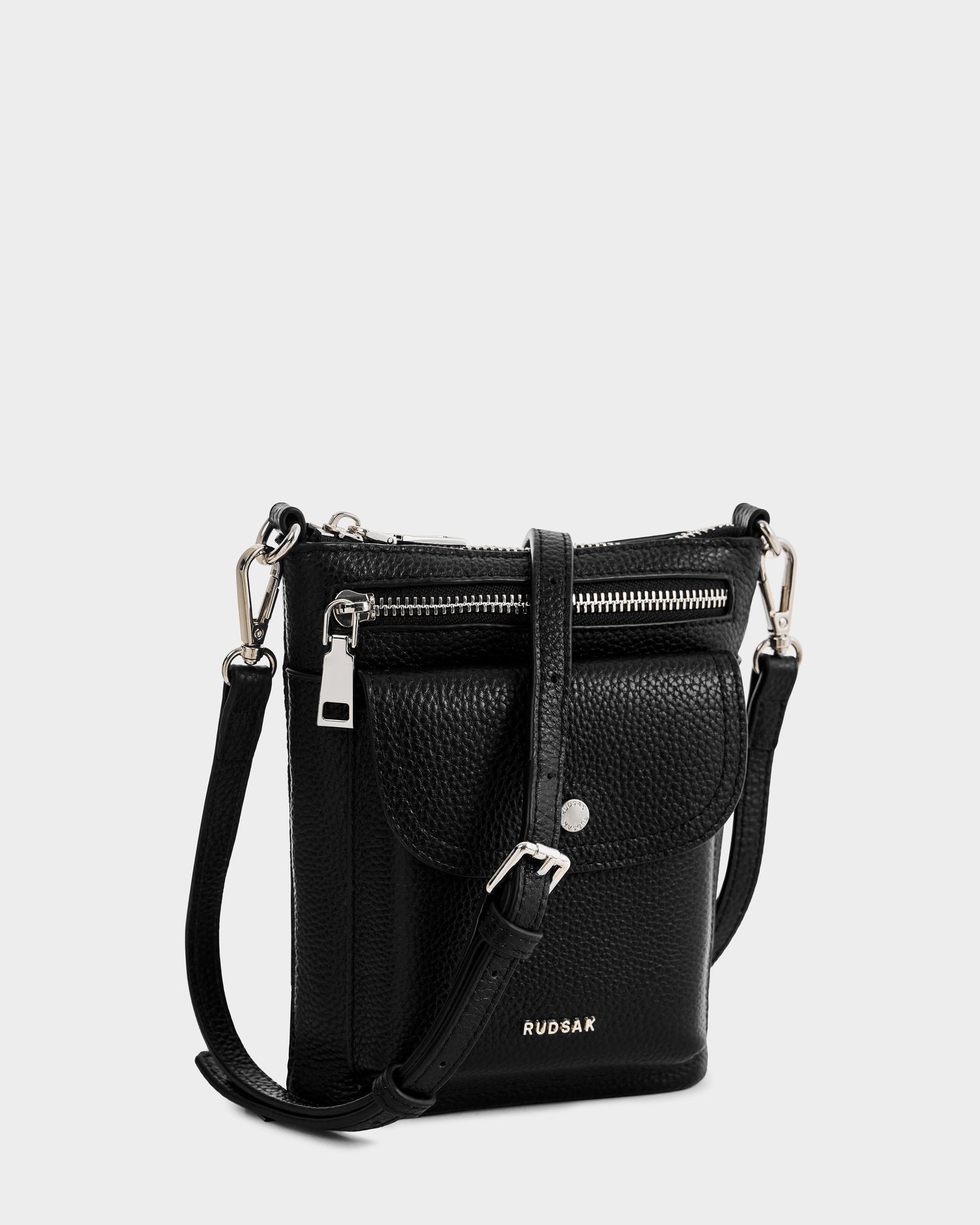 The LILA - L325513 Black by Rudsak is a small black women’s crossbody bag in pebbled leather, featuring a front flap pocket, silver zipper, adjustable strap, and "RUDSAK" printed in silver on the front.