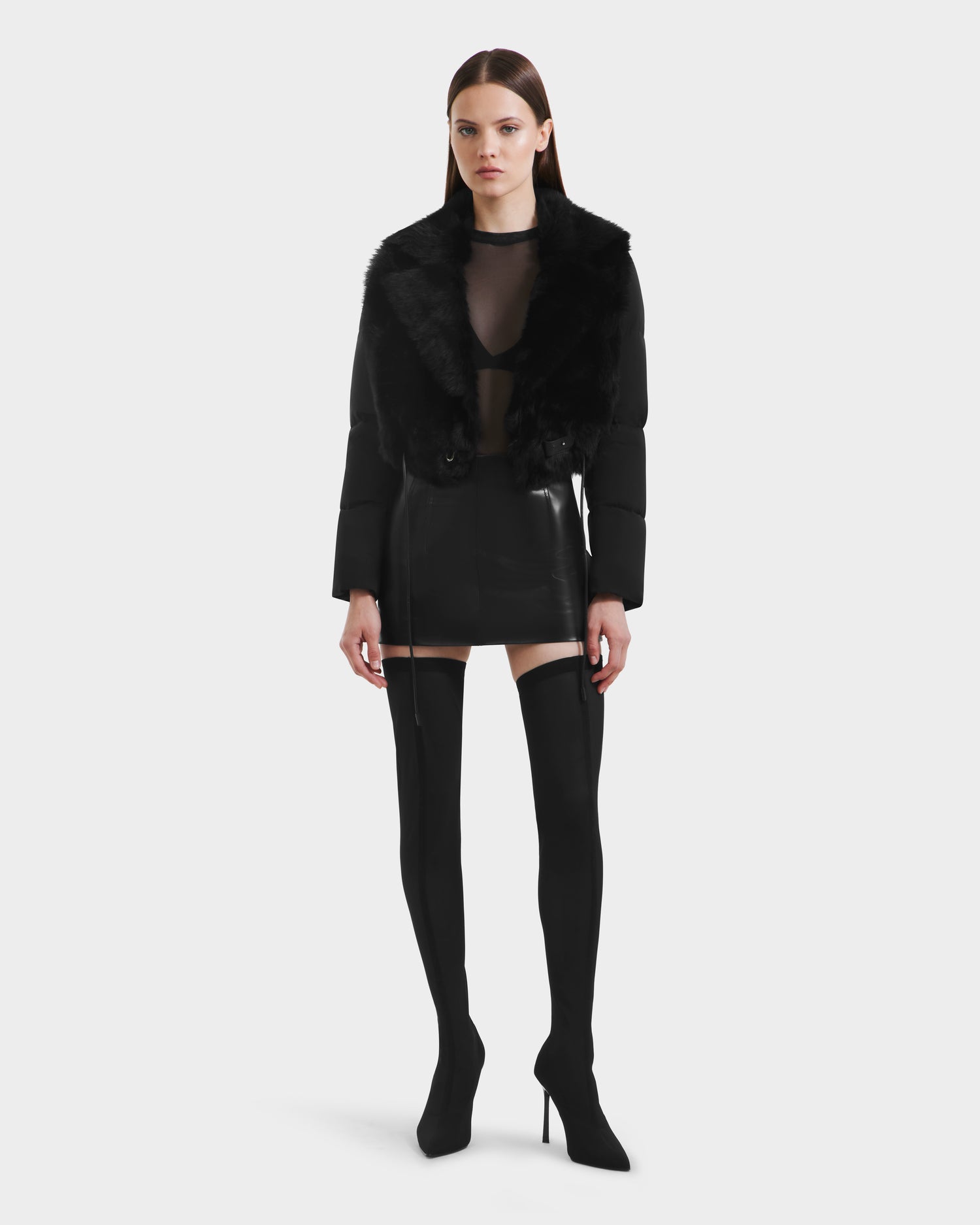 A woman poses against a plain white background wearing Rudsak’s LILAH - 8125509 Black shearling lamb jacket paired with a sheer black top, a black leather mini skirt, and thigh-high black stiletto boots.