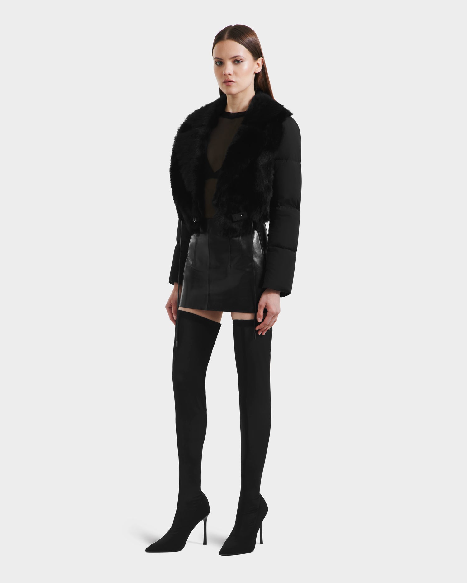 A woman in the Rudsak LILAH - 8125509 Black women’s cropped jacket with a faux fur collar, sheer black top, mini skirt, and thigh-high heeled boots stands against a white background.