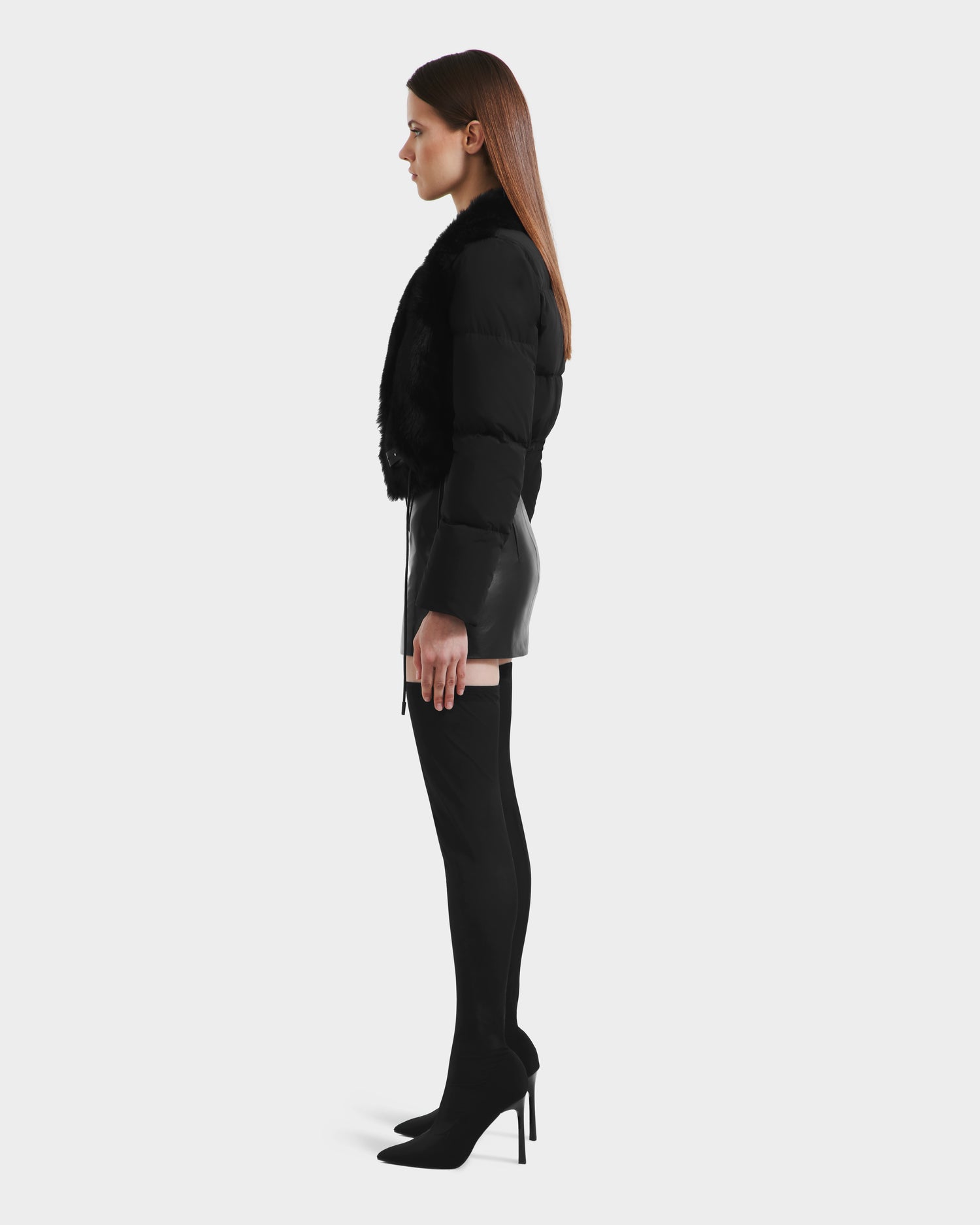 A woman stands in profile against a plain background, wearing the Rudsak LILAH - 8125509 Black women’s cropped quilted puffer jacket with fur trim, a black leather mini skirt, black thigh-high boots, and black high heels.